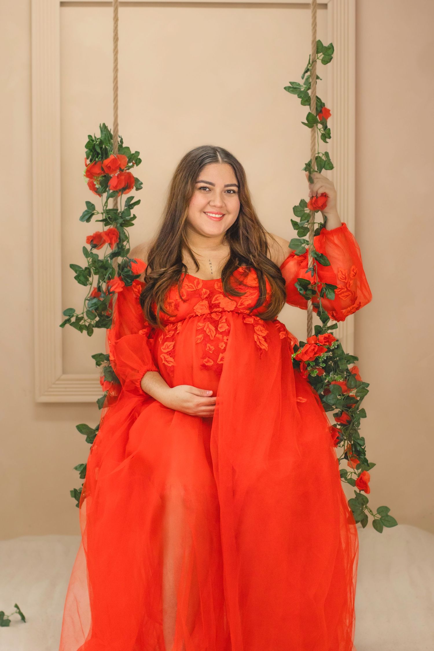 A flowing red sheer maternity gown with floral decorations and green vines against a beige wall.