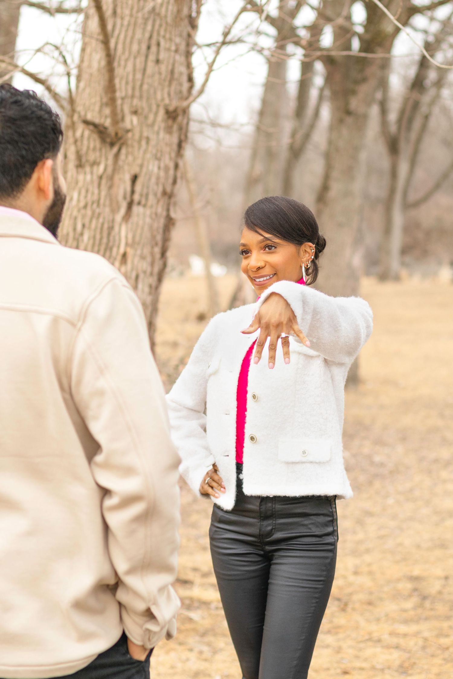 A woman in a white jacket and pink scarf shows off her engagement ring outdoors.