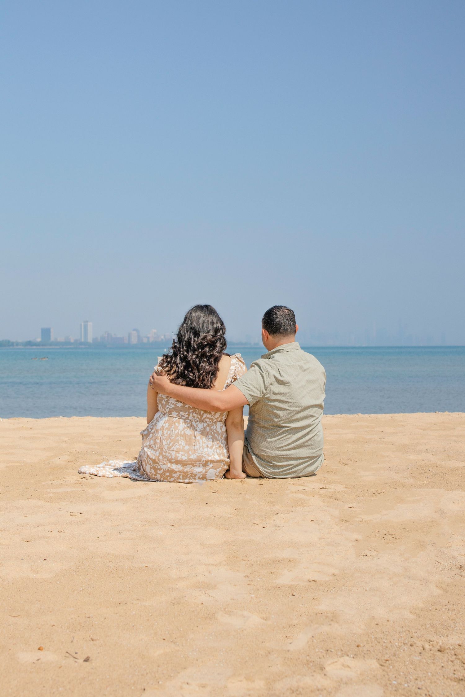 Couple sitting together on sandy beach looking at city skyline across calm blue waters on sunny day.