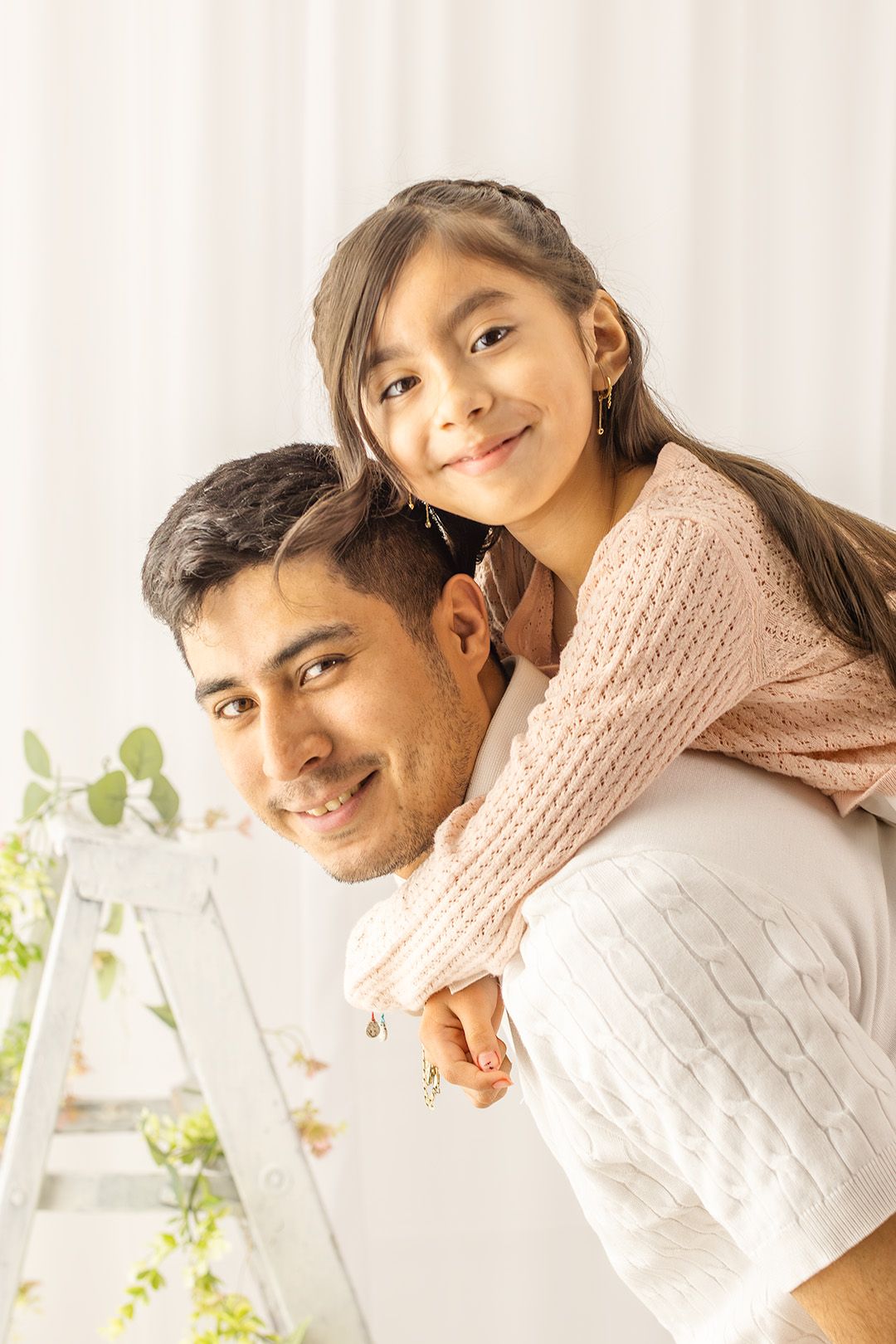 A man gives a young girl a piggyback ride, both smiling warmly in a bright, cozy indoor setting.