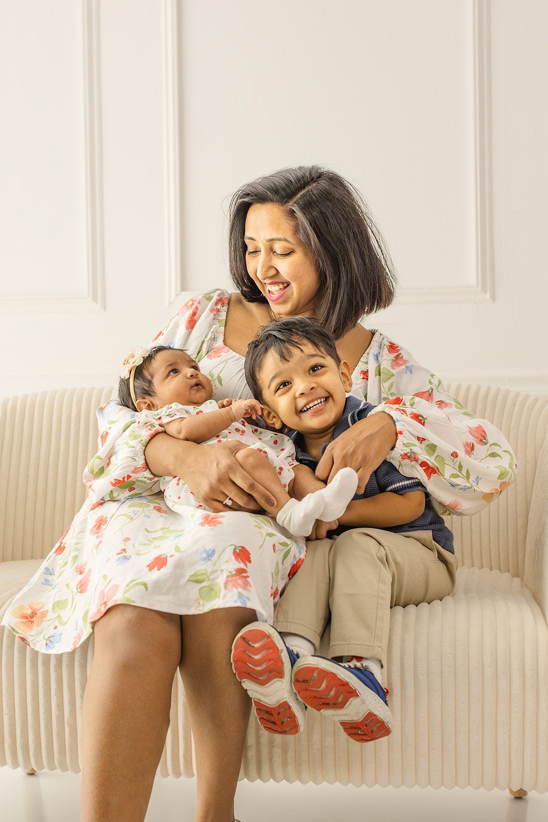 A smiling mother in a floral dress sits on a sofa holding her baby girl and young son, sharing a joyful family moment.