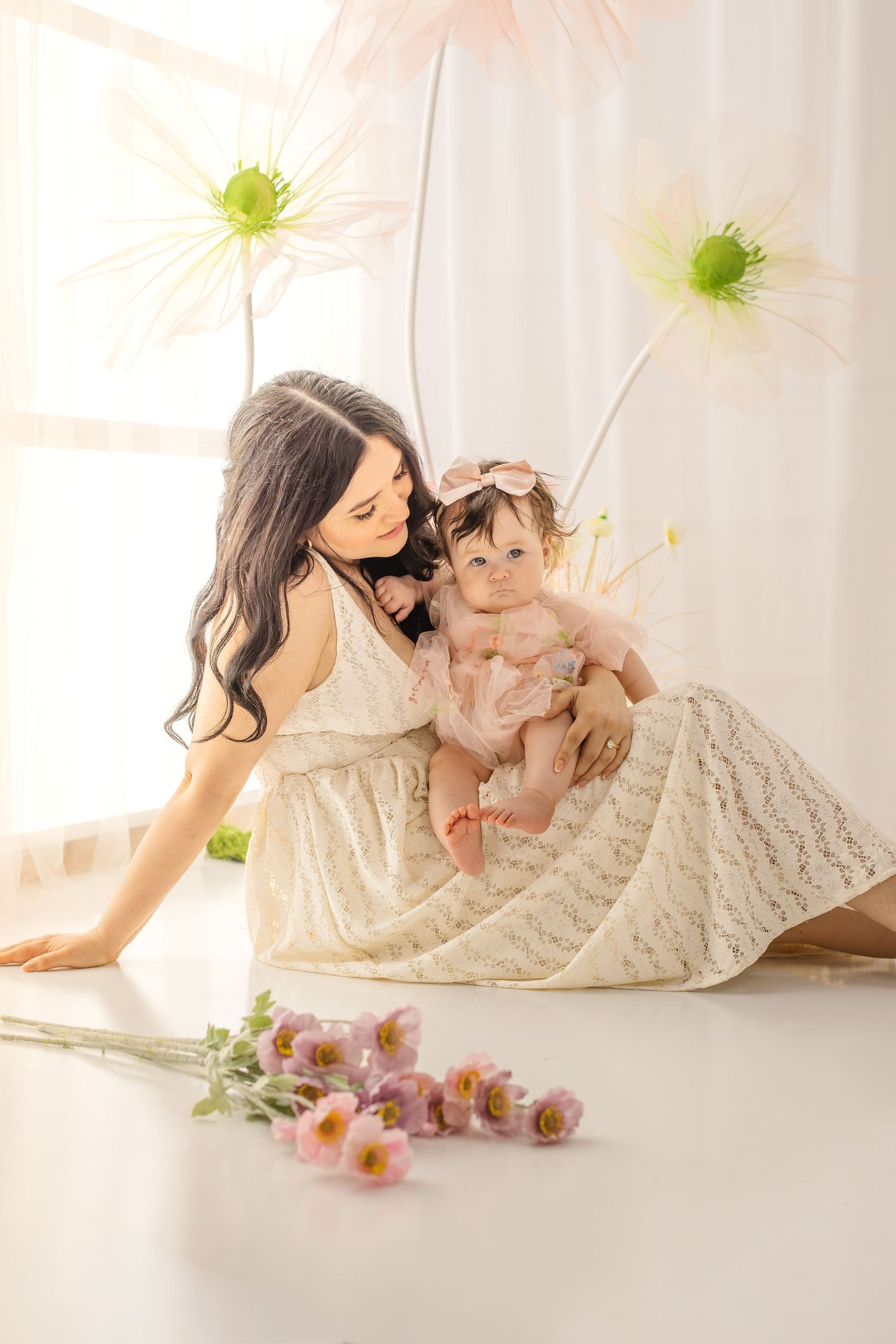 A mother in a white lace dress tenderly holds her baby girl in a pink tutu amid large floral decorations.