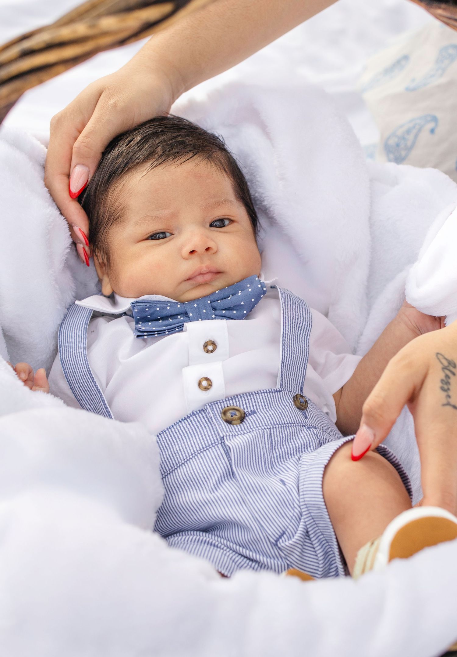 A baby in a blue bow tie and suspender outfit lies on white blankets looking dapper and adorable.