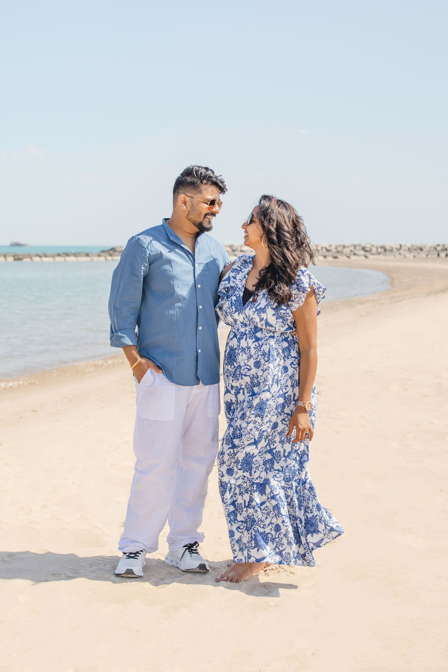 Two people standing together on a sunny beach, with one wearing a blue vest and white pants and another in a floral dress.