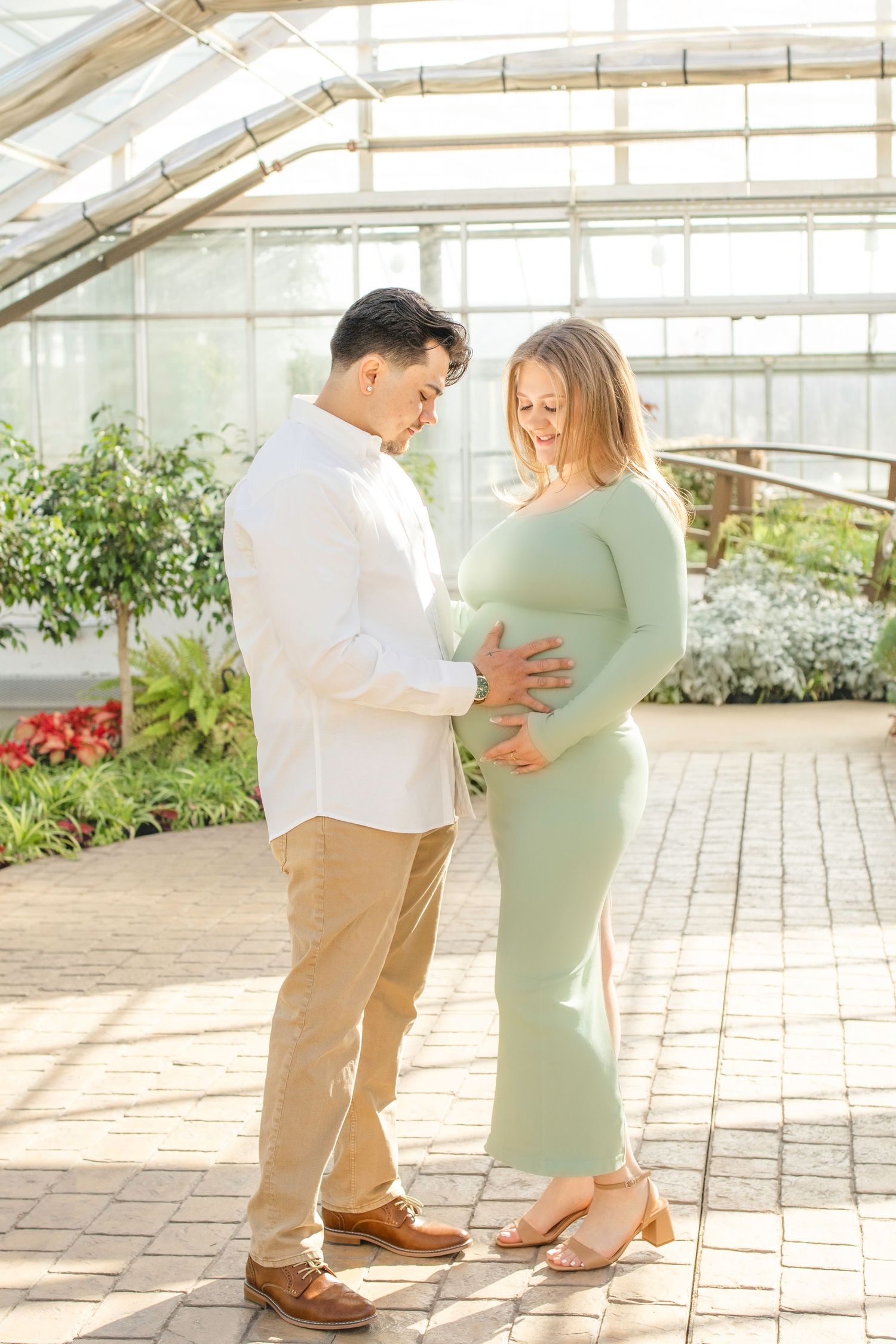 Expectant couple shares tender moment in sunlit greenhouse surrounded by blooming plants and flowers.