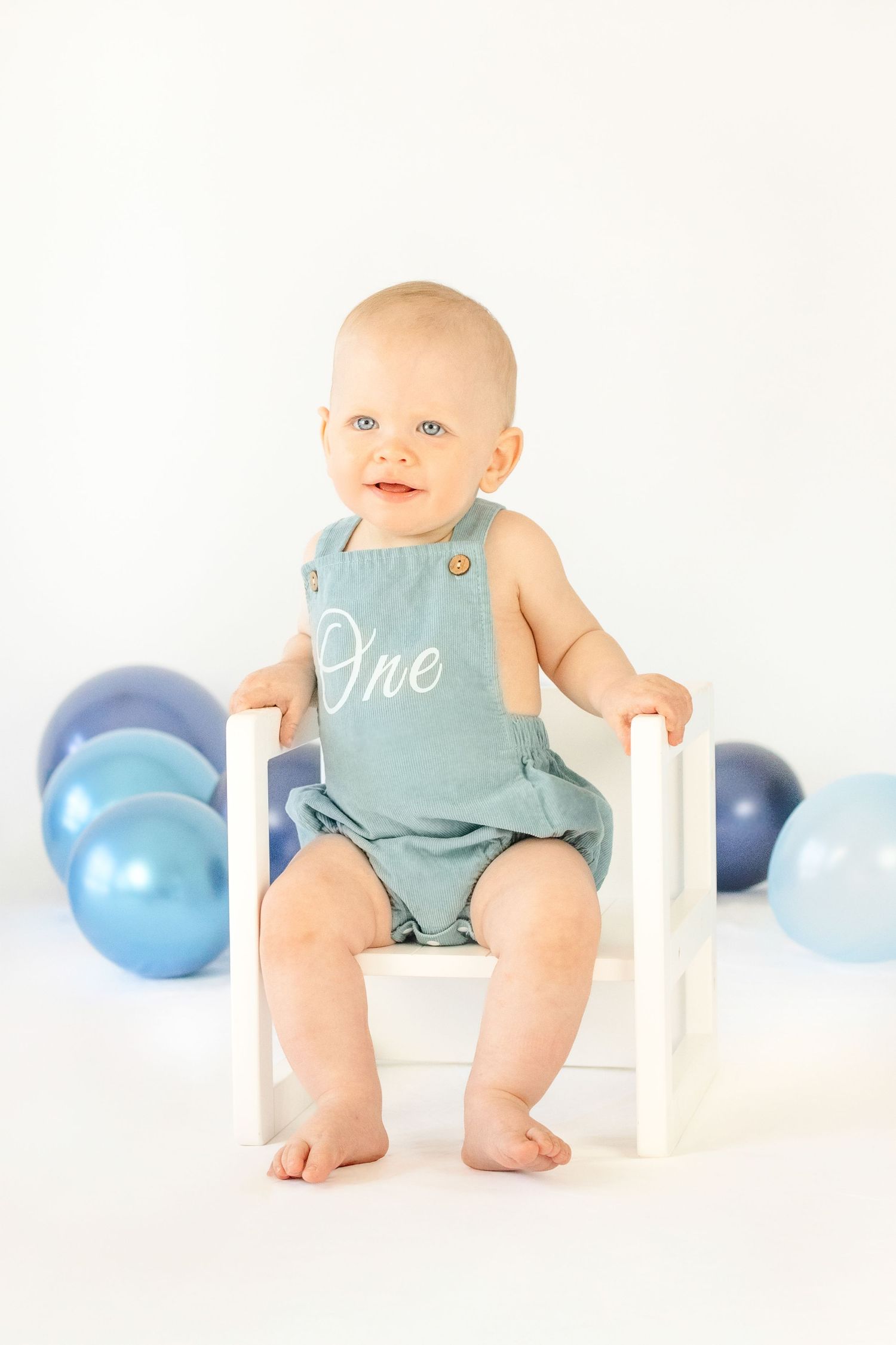 Baby in blue “One” romper sits on white chair surrounded by blue balloons celebrating first birthday.
