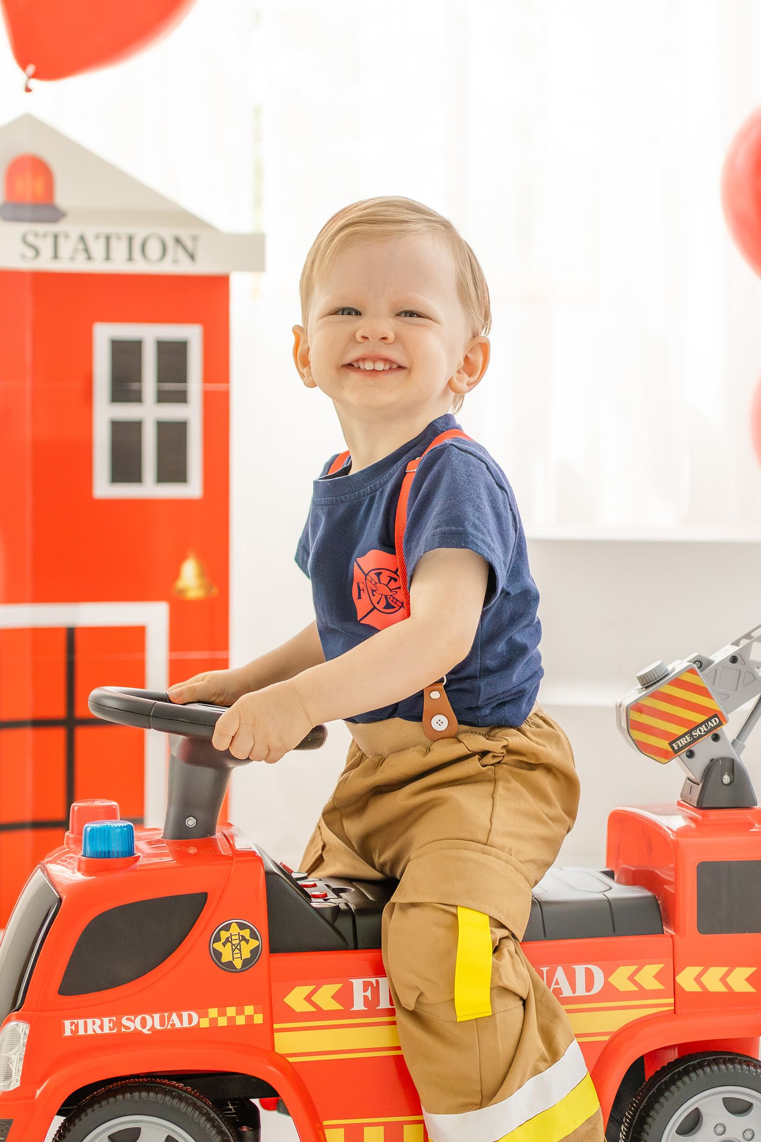 Smiling toddler boy in firefighter costume riding red toy fire truck at birthday party.