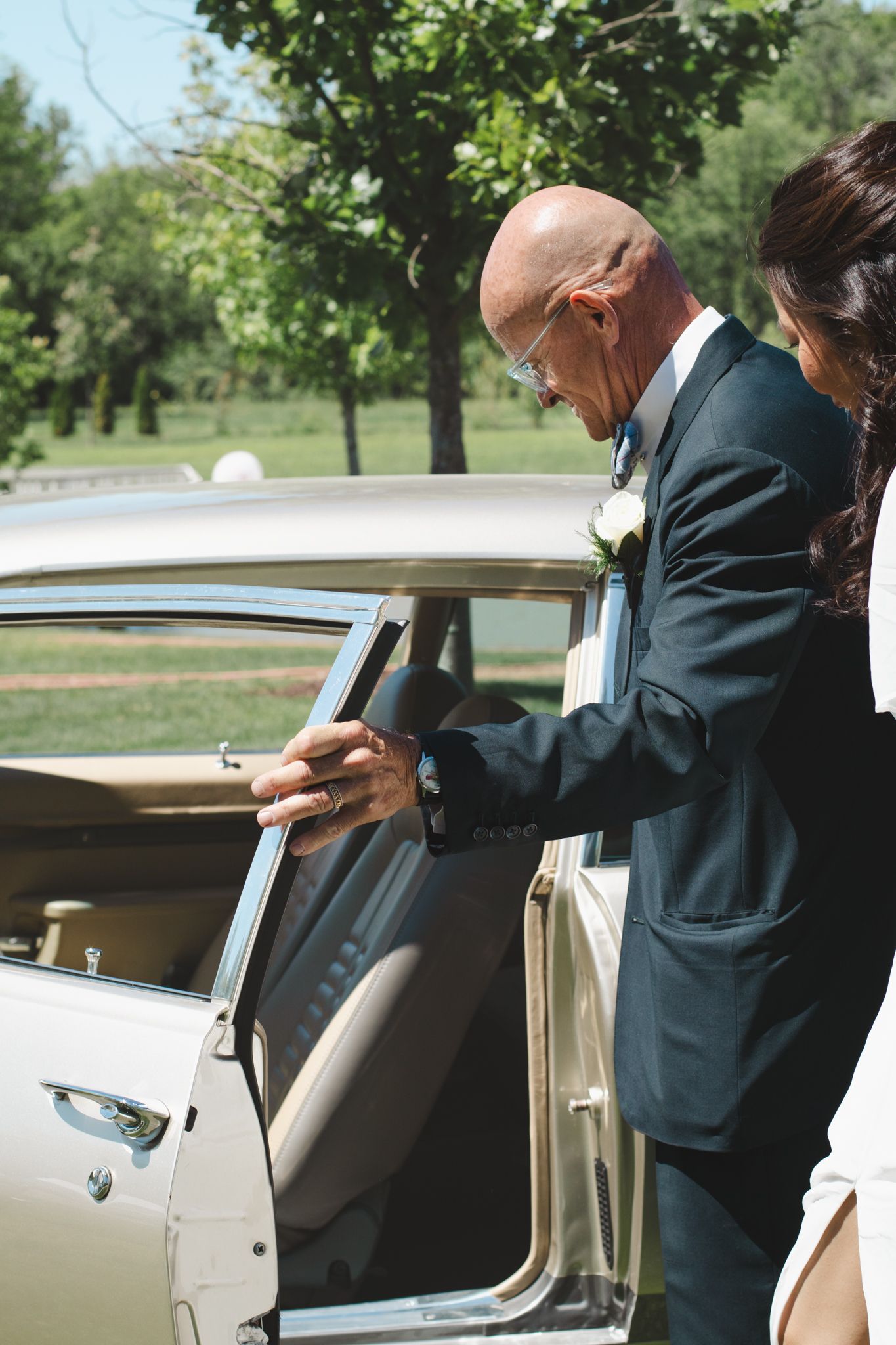 Wedding scene showing attendant opening vintage car door for couple on sunny outdoor day.