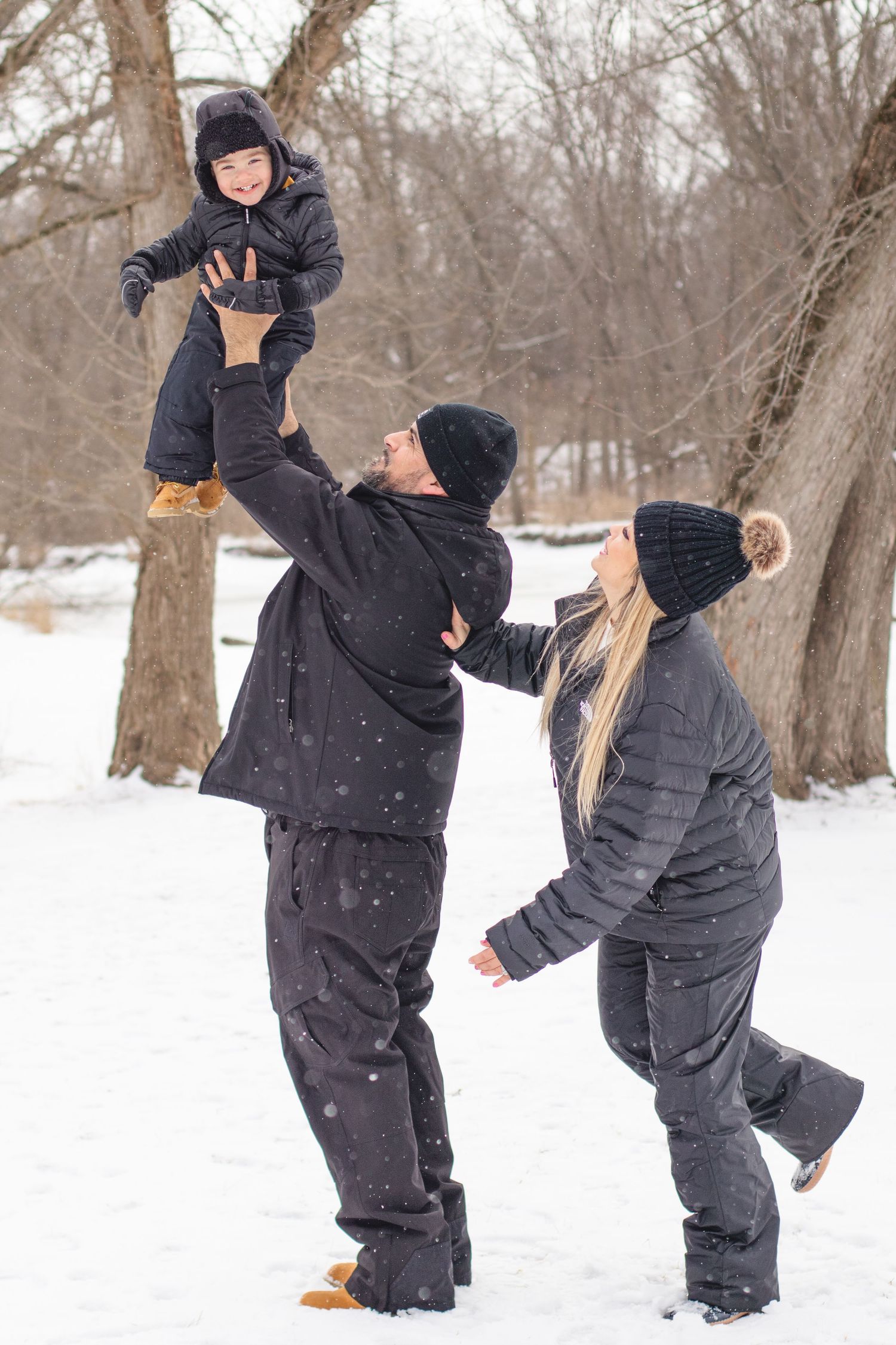Parents play with their child in winter snow, lifting them up playfully among bare trees.