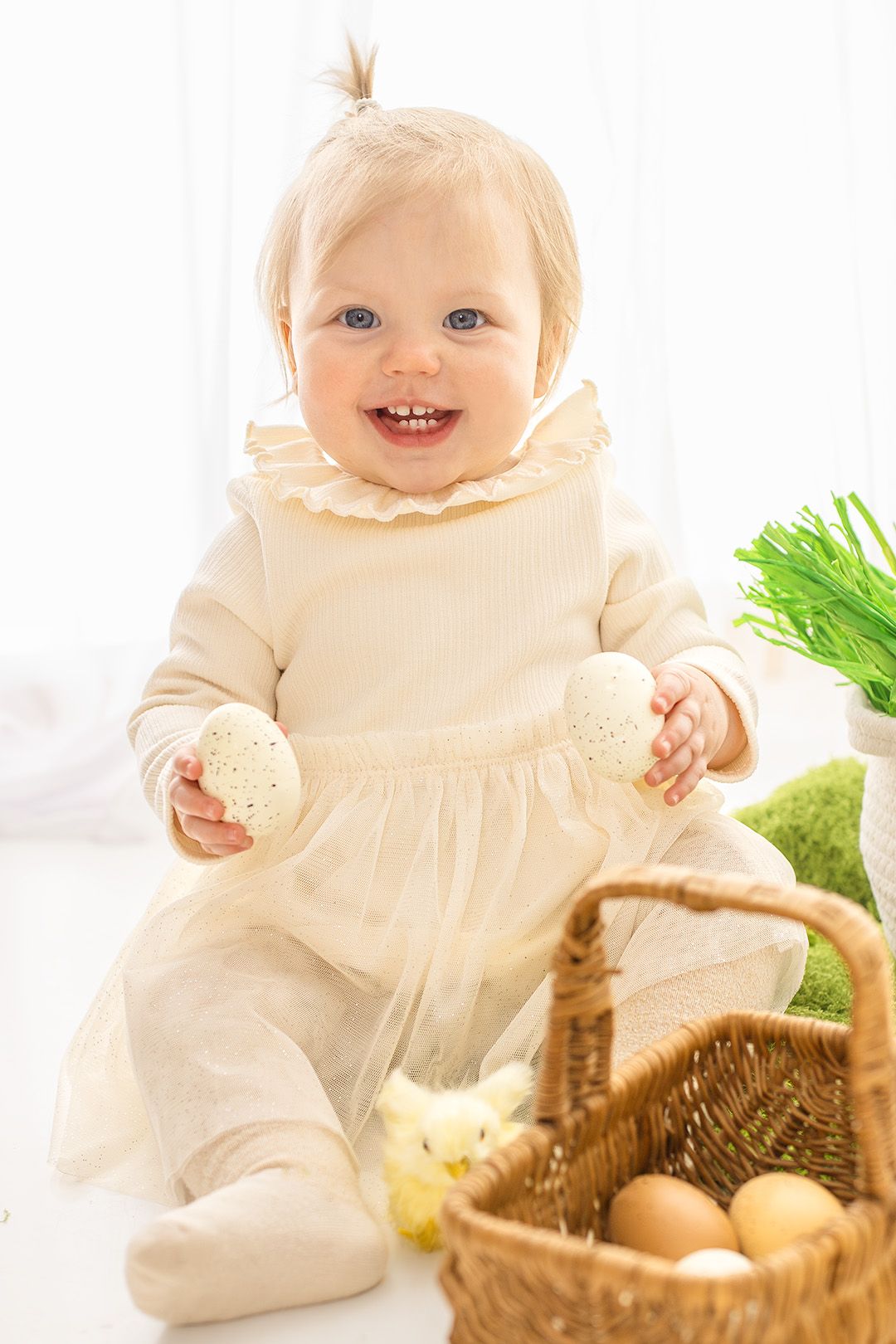 Baby in cream-colored dress sits near Easter basket with eggs and spring decorations.