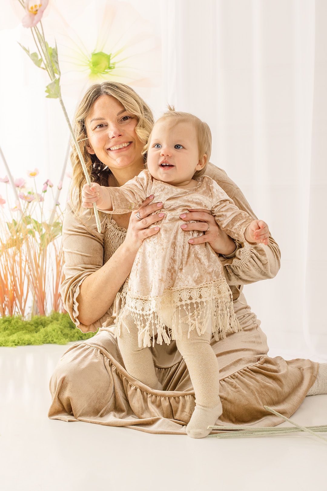 A parent and toddler wearing matching beige outfits pose together in a bright, airy setting with floral decorations.