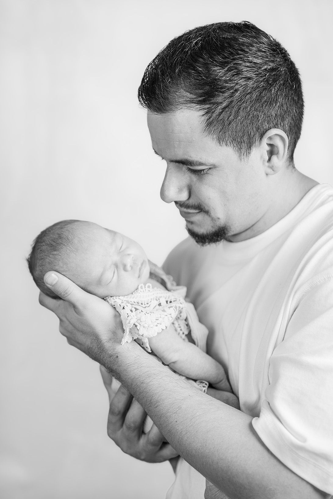 Black and white photo of a young father tenderly cradling his sleeping newborn baby in both hands.