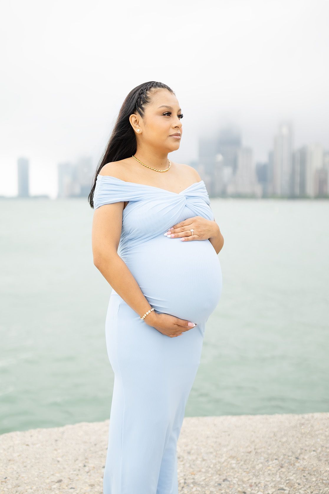 Pregnant woman in light blue off-shoulder gown cradles baby bump by waterfront with city skyline backdrop.