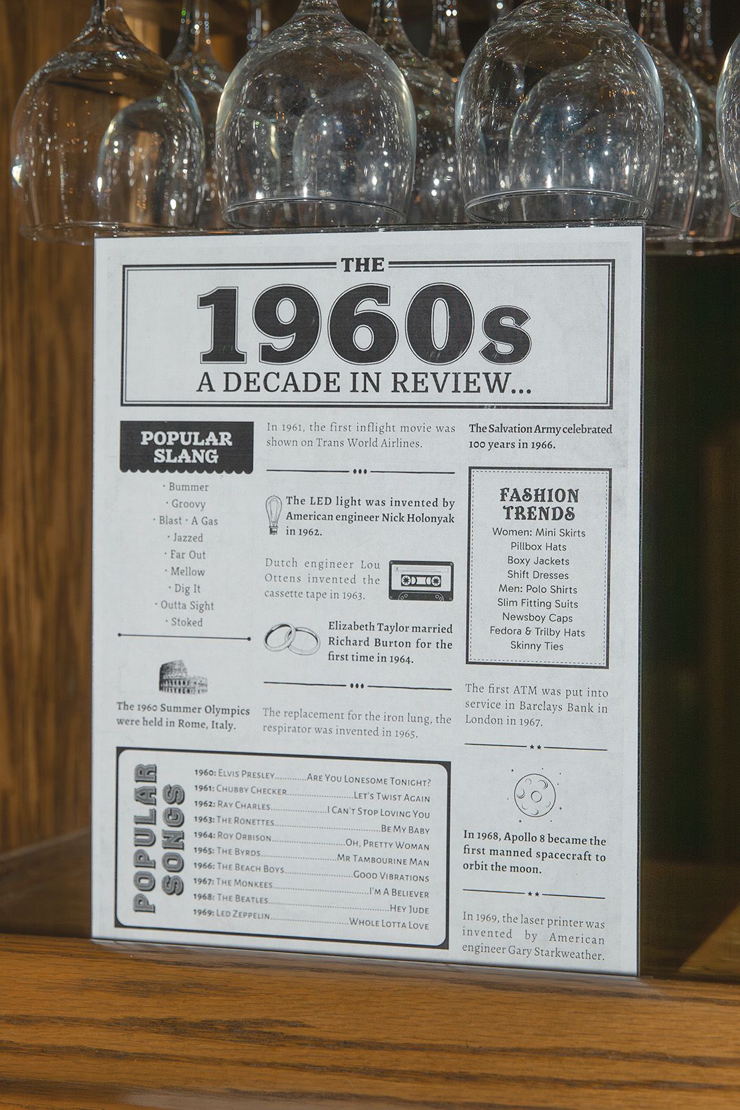 A vintage-style 1960s decade in review informational poster displayed on a wooden surface at a party.