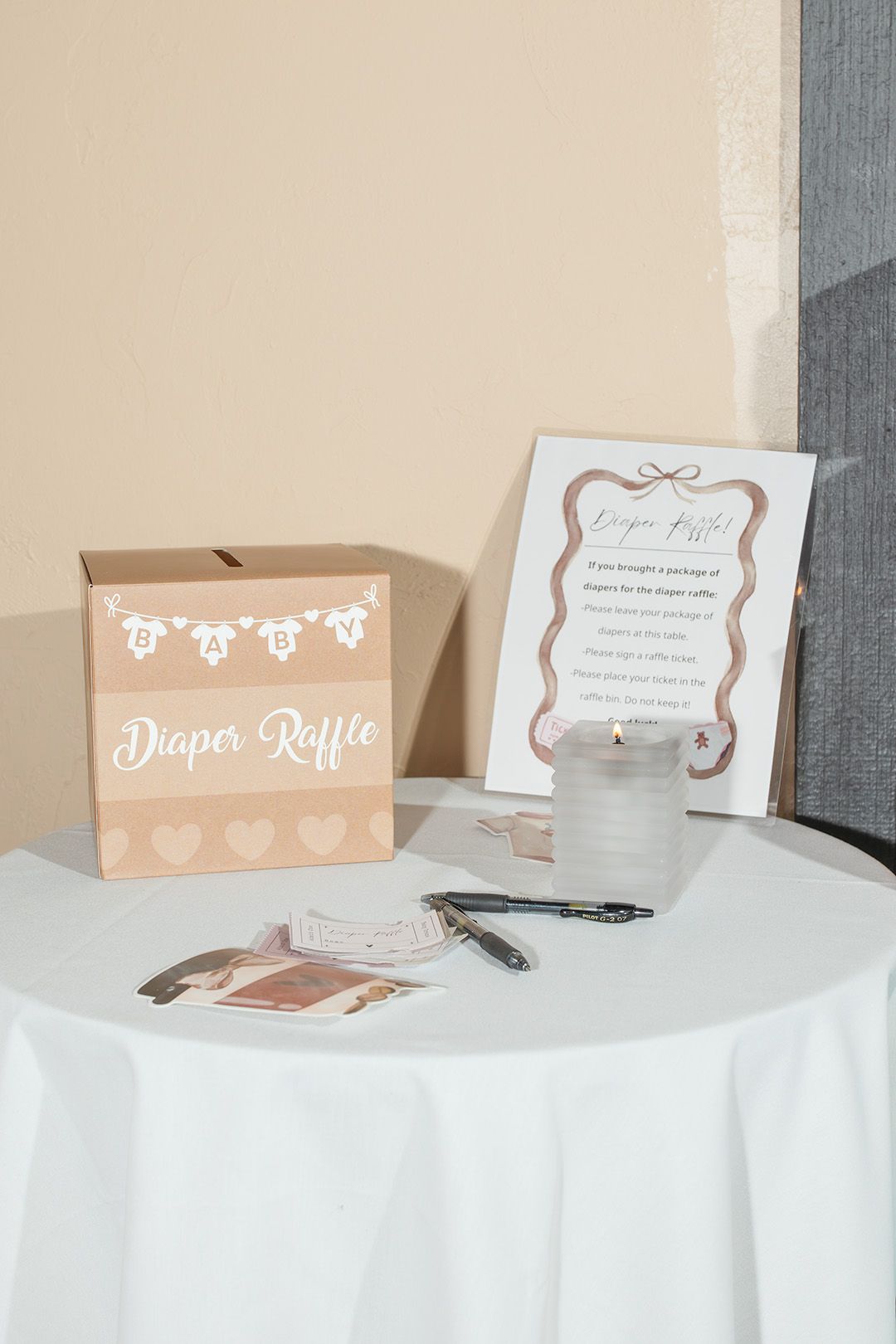 Baby shower diaper raffle table with kraft paper box, sign, candle, and pens on white tablecloth.
