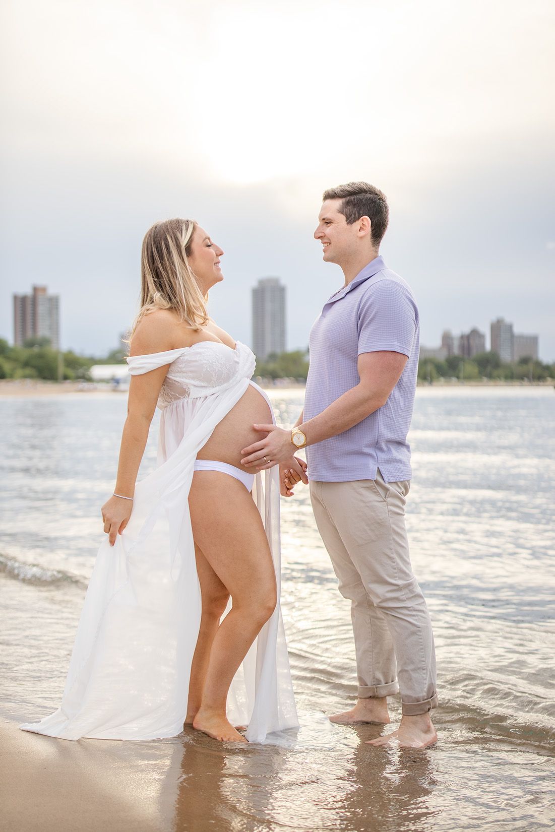 Pregnant couple holding hands on a sunny beach, woman in white dress cradling baby bump, city skyline in background.
