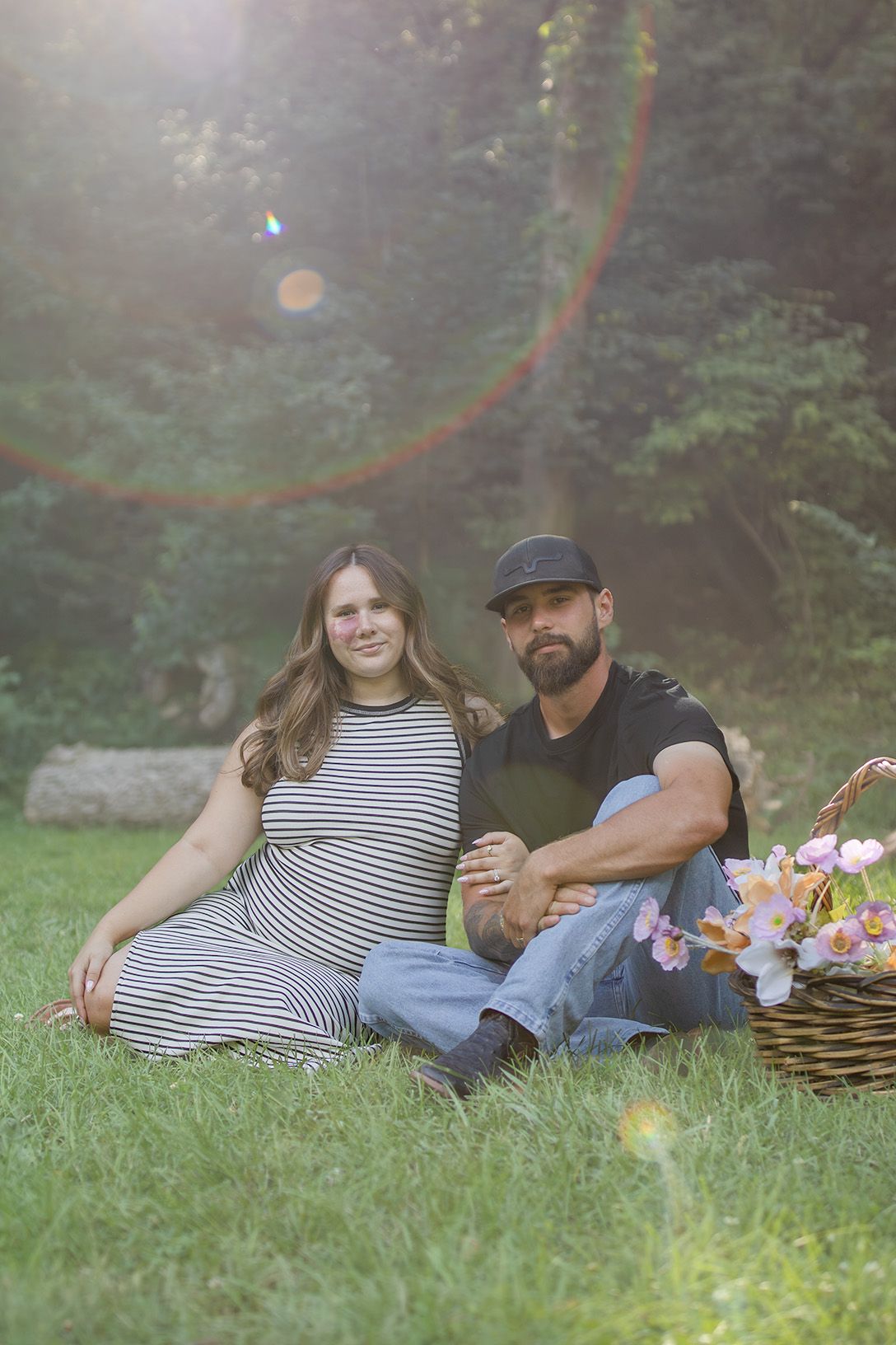 Smiling couple sitting on grass with flower basket, pregnant woman in striped dress with partner in cap.
