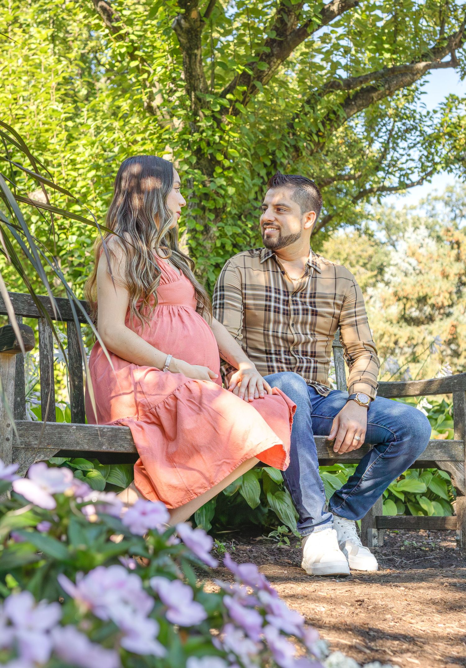 Couple relaxing on a garden bench surrounded by purple flowers and lush greenery on a sunny afternoon.