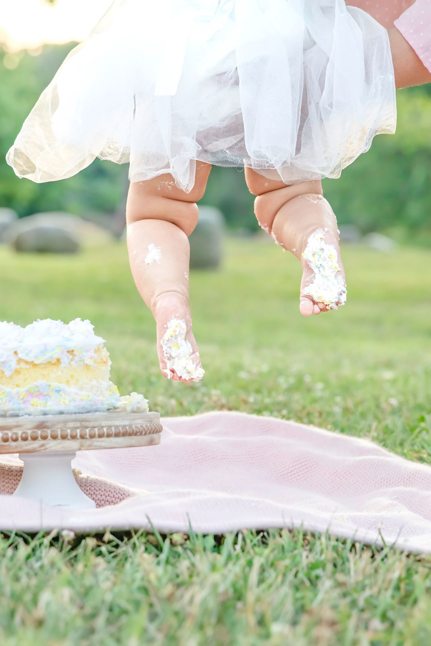 Baby feet covered in cake frosting dangle above a picnic blanket with a smashed cake during first birthday photoshoot.
