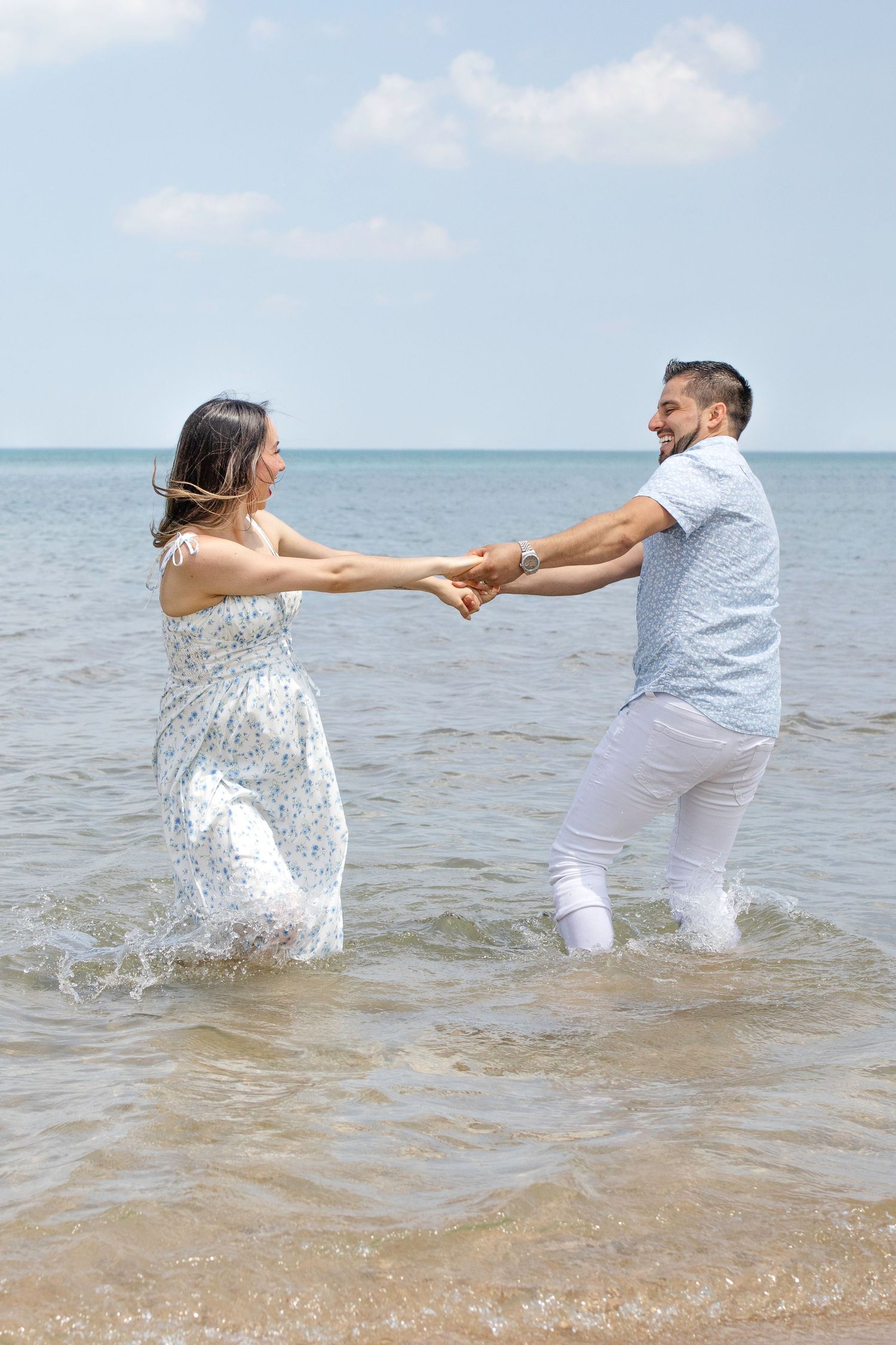 A couple in white holds hands while playing and splashing in shallow ocean water during a beach photoshoot.