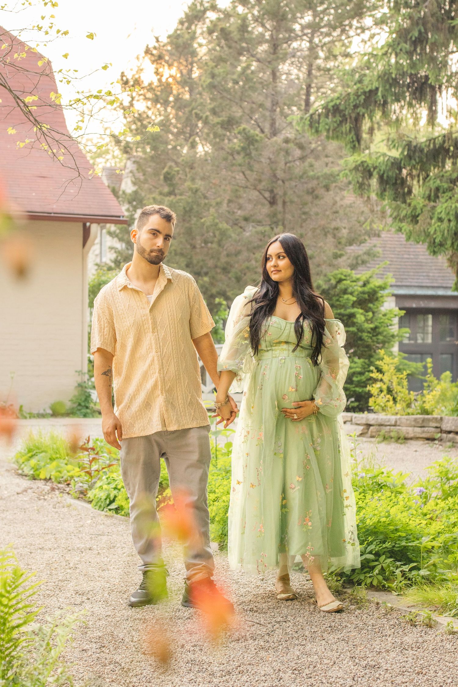 A couple in coordinated earth-tone outfits walking together on a gravel path surrounded by lush summer greenery.