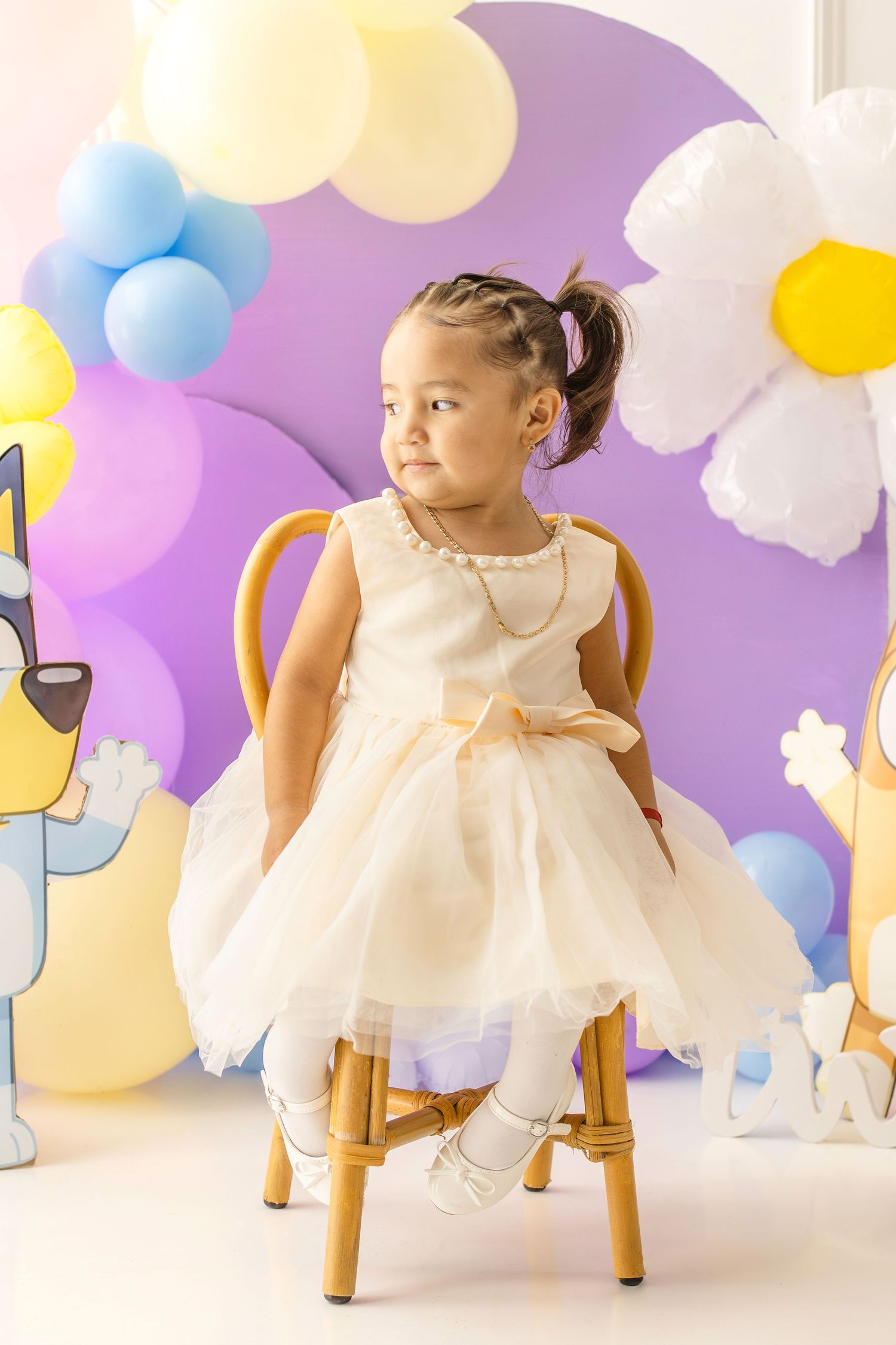 Toddler girl in cream tulle dress sits on wooden chair at birthday party with purple backdrop and colorful balloons.