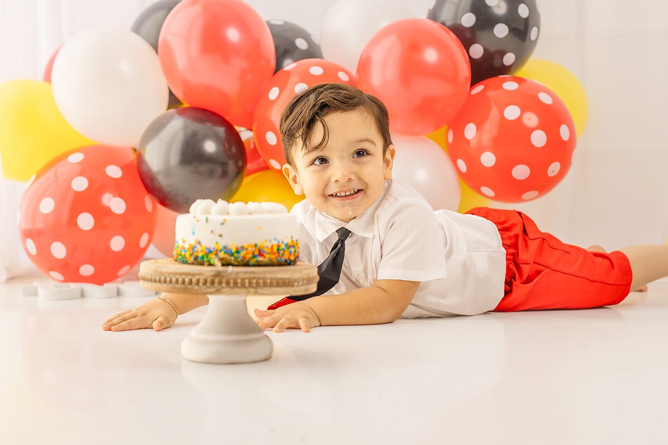 Smiling toddler boy in red pants and tie poses with birthday cake surrounded by colorful Mickey Mouse balloons.