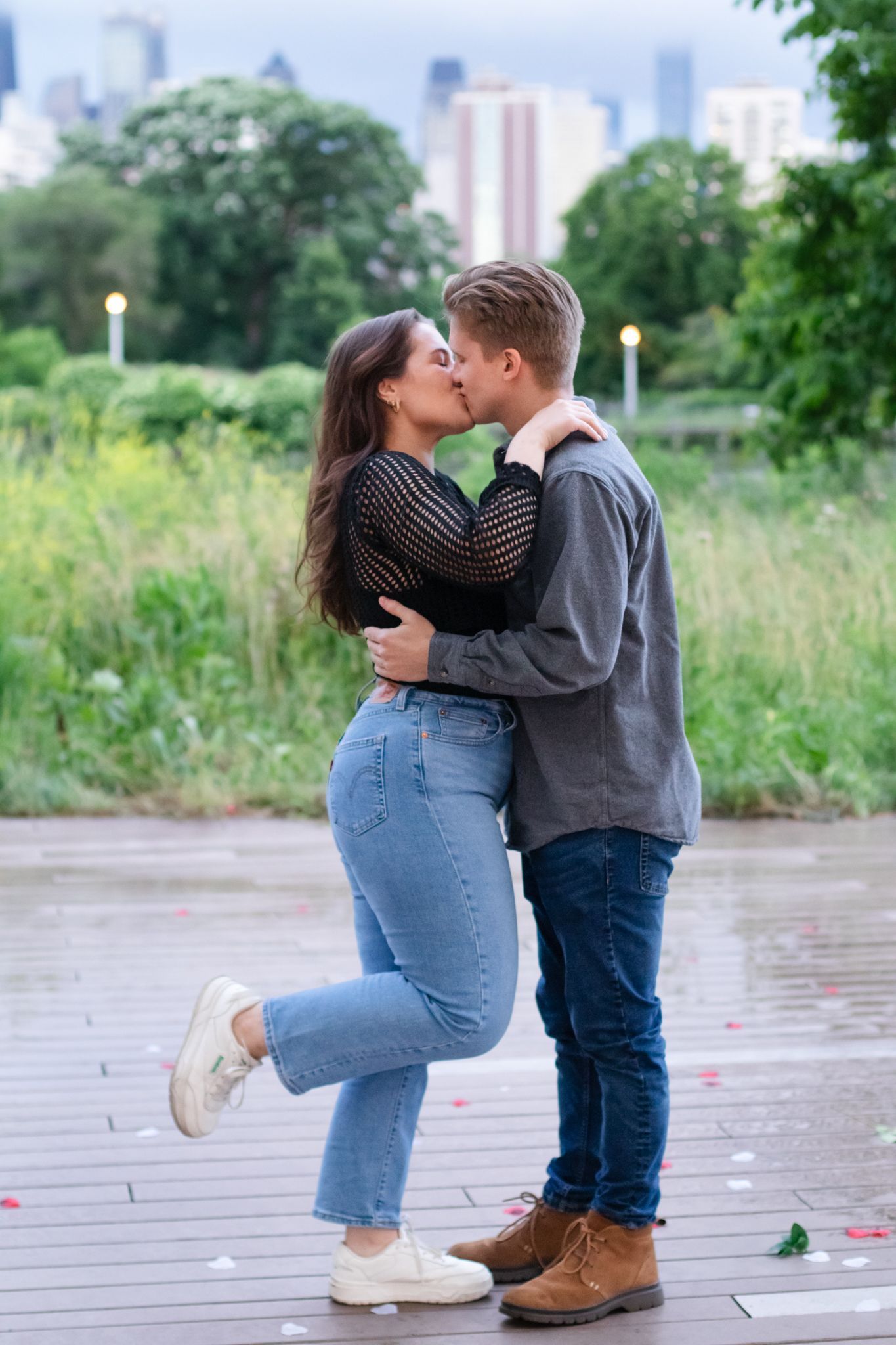 Couple shares romantic kiss on wooden deck with city skyline in background.