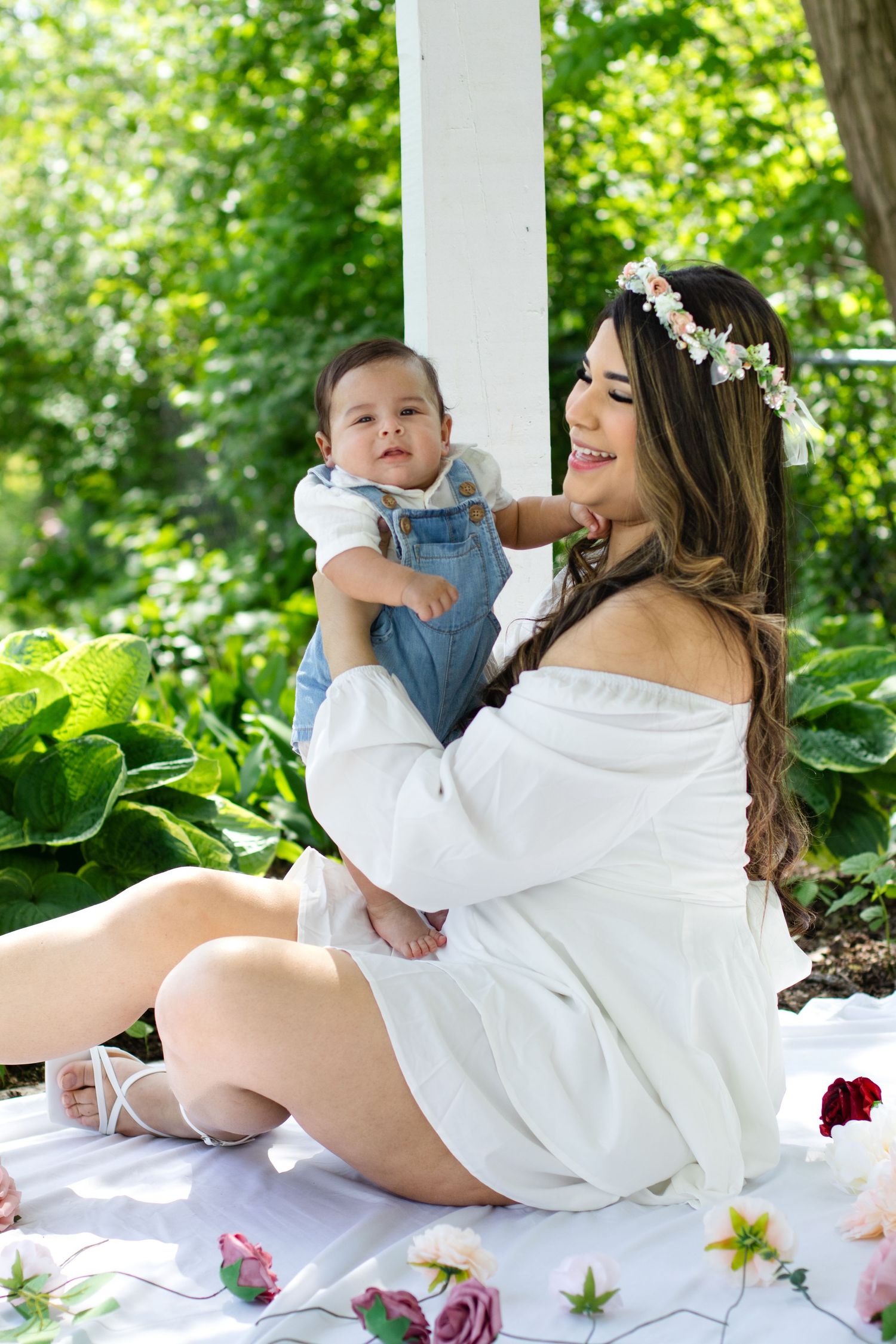 Mother and baby in white dress have springtime photoshoot on blanket with flowers.