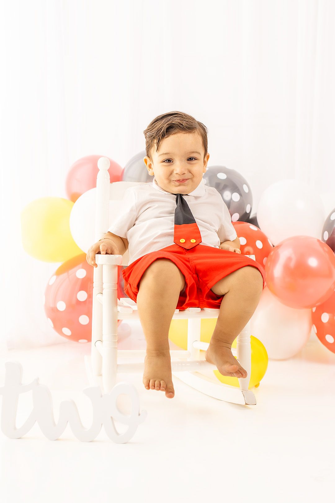 Toddler boy in Mickey Mouse outfit sits on white chair surrounded by colorful balloons at second birthday party.