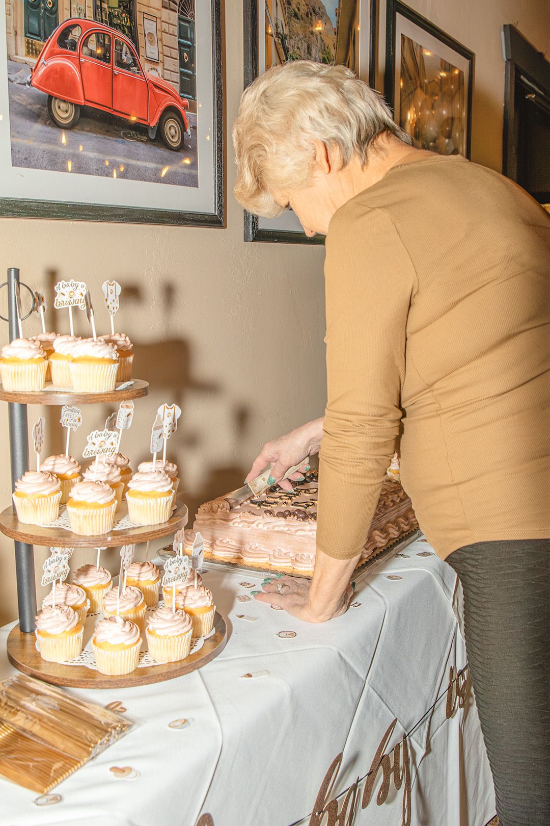 Elderly woman cutting a decorated cake at a party table with tiered cupcake stand and framed art on wall.