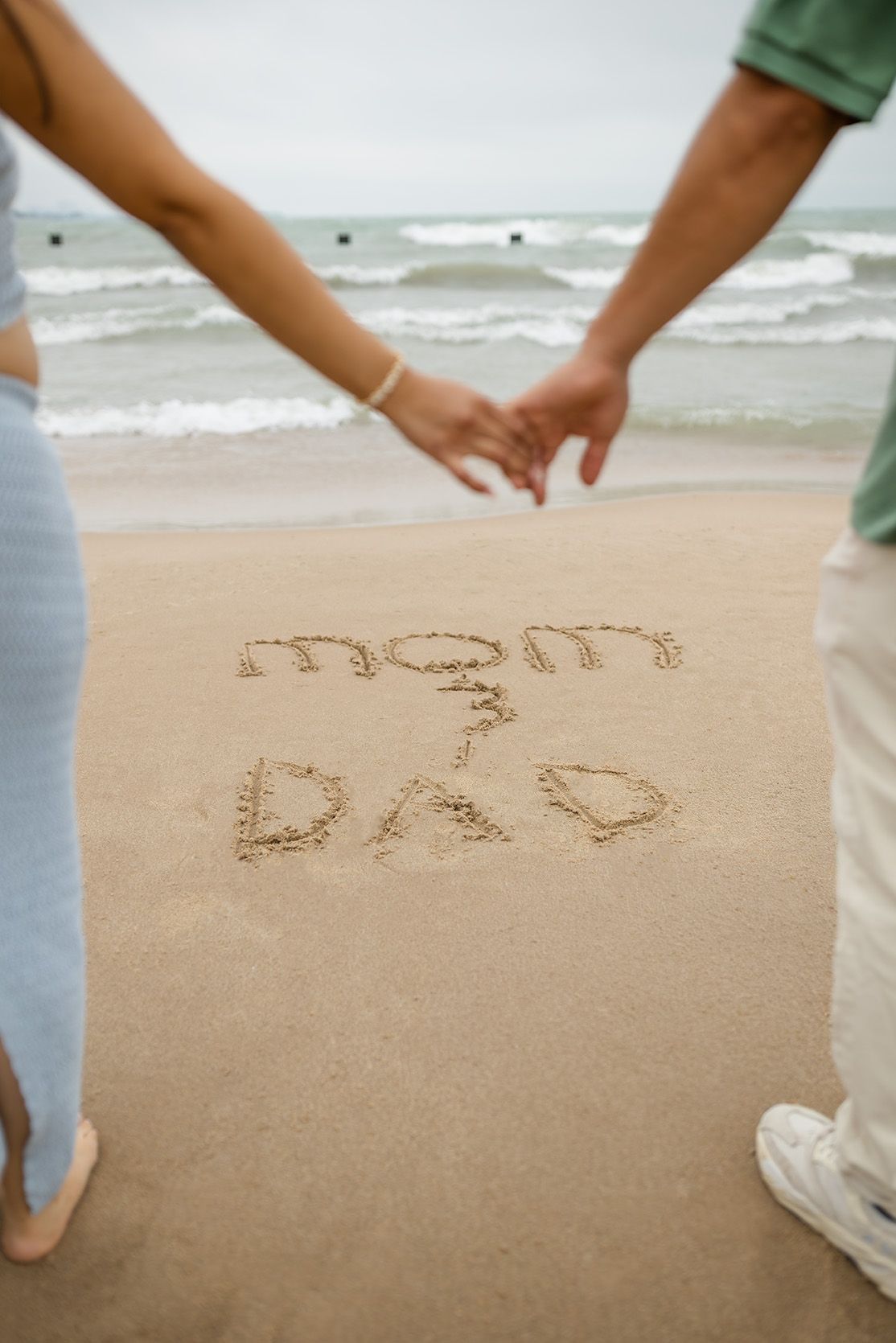 Couple holding hands on beach with mom and dad written in sand near ocean waves.