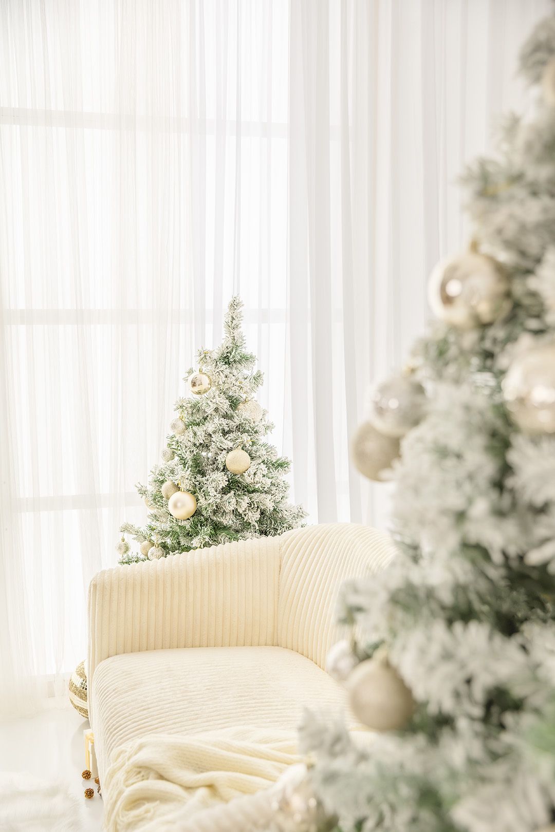 Elegant white Christmas room with flocked trees, gold ornaments, and a cream armchair by sheer curtains.