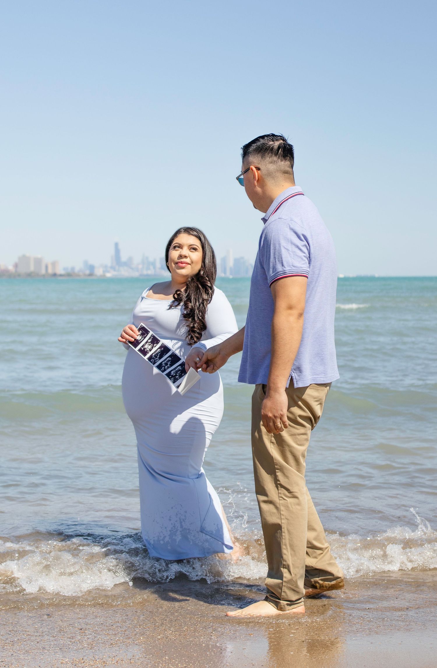 A couple stands at the edge of Lake Michigan in Chicago, with the woman wearing a white dress for maternity photos.