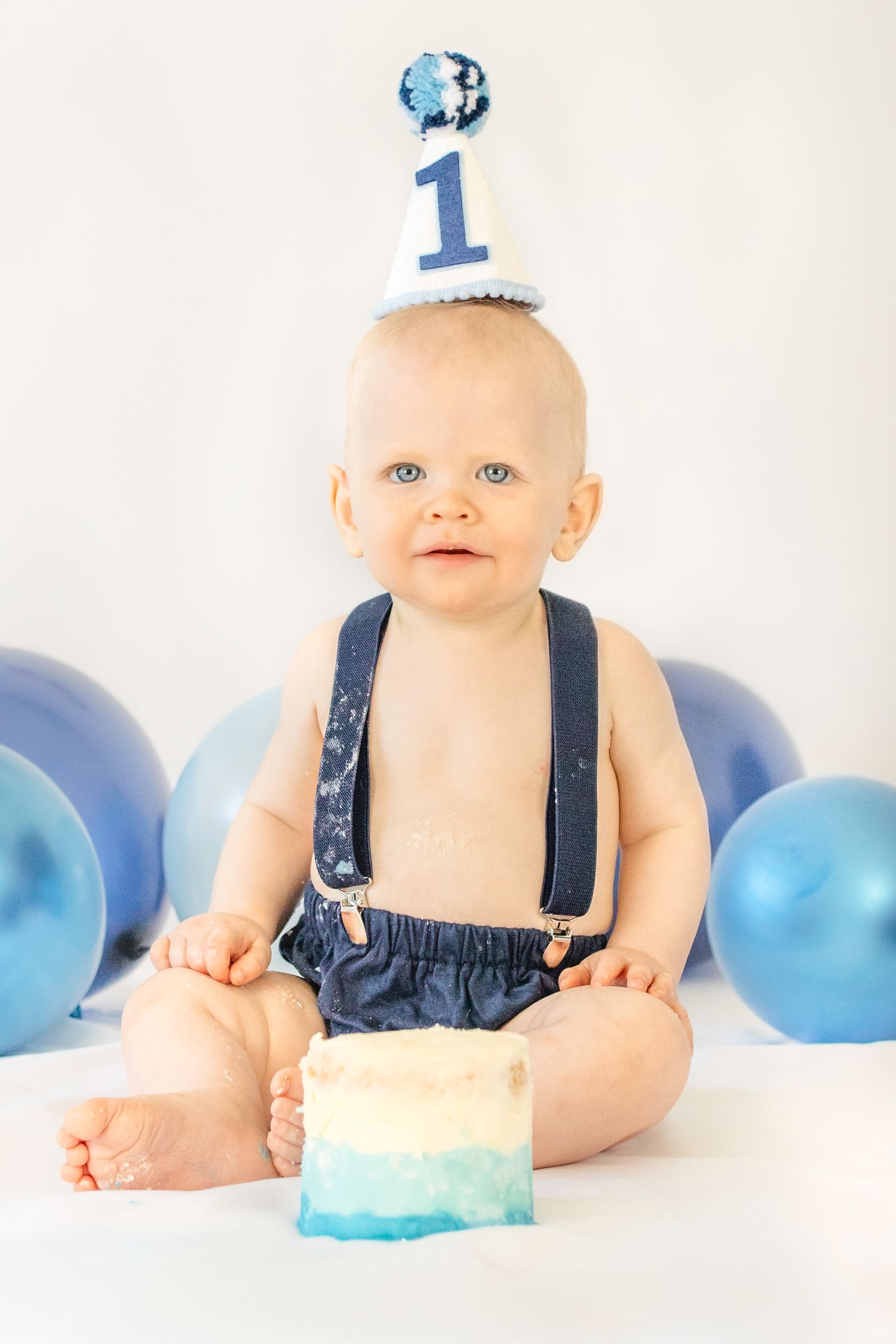 Baby boy in blue suspenders and birthday hat sits with smash cake and balloons celebrating his first birthday.