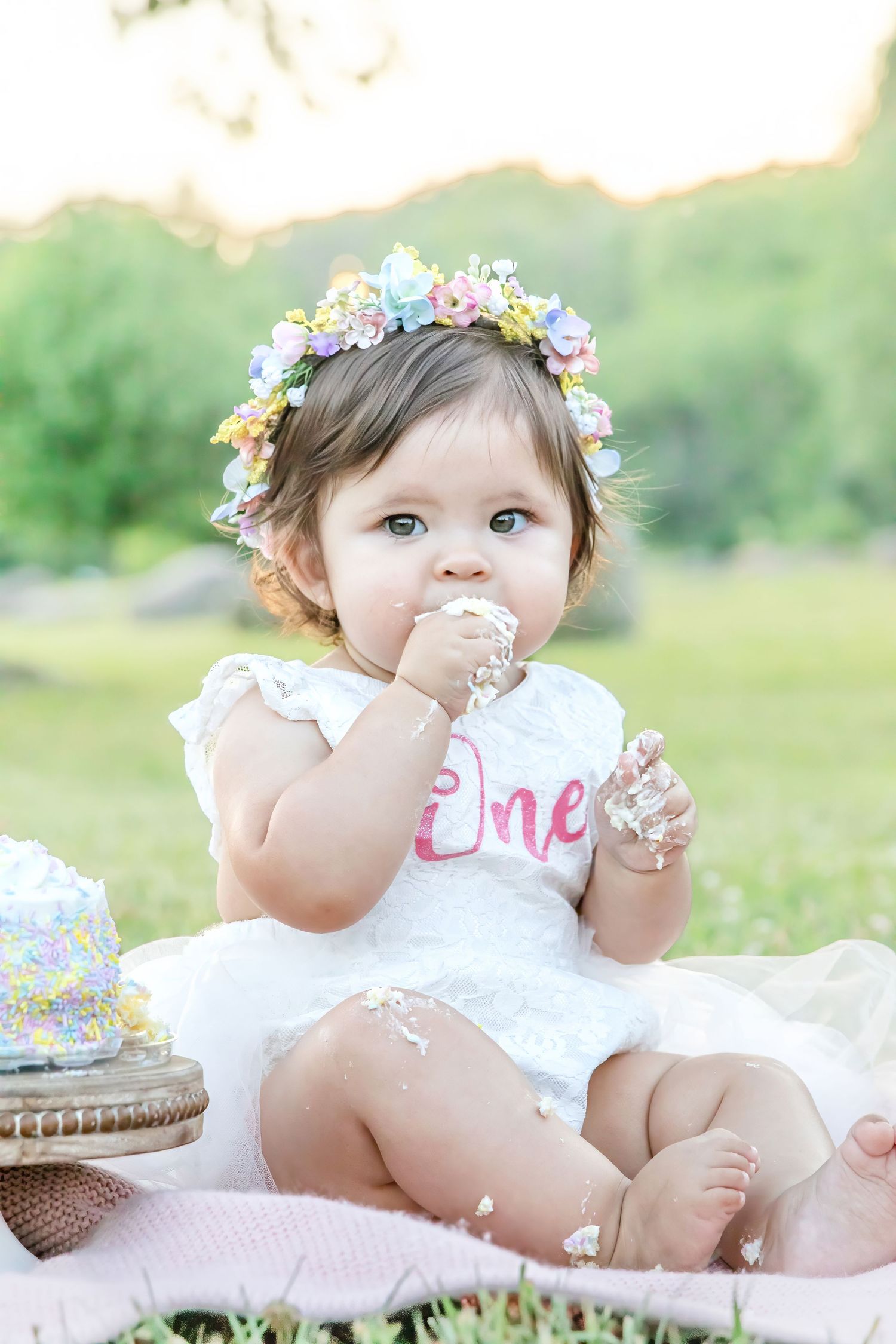 Baby girl in floral crown eating cake at outdoor first birthday smash cake session wearing white lace romper.