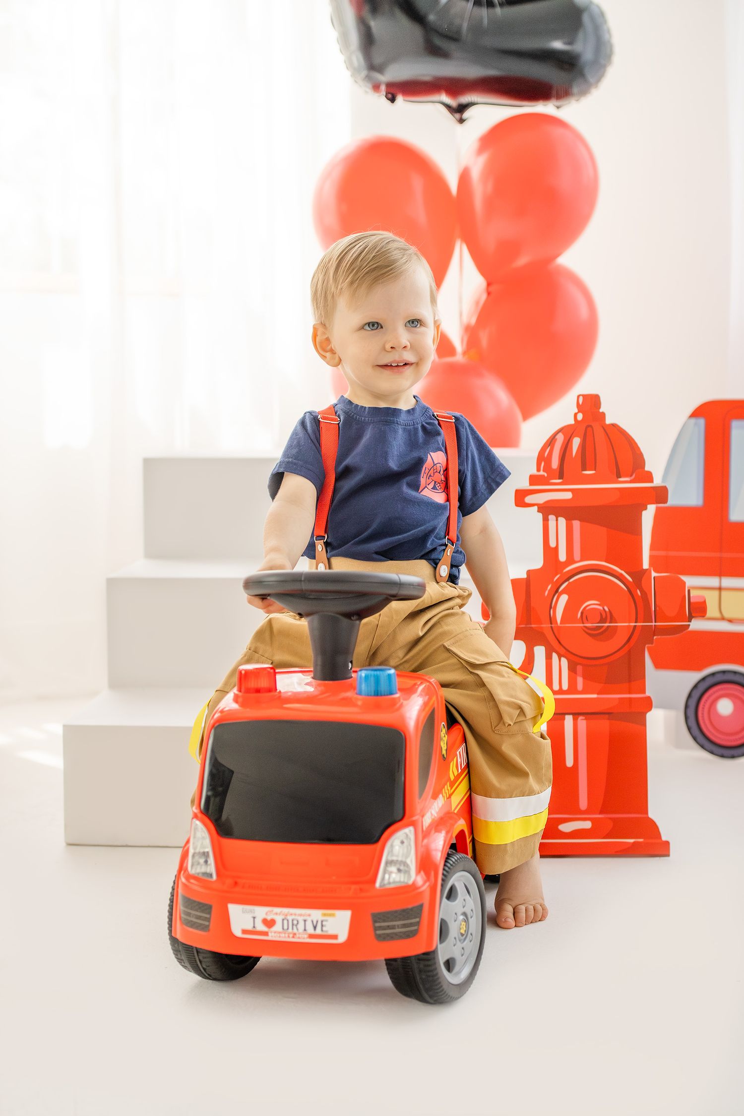 Happy toddler in firefighter outfit sitting on red ride-on toy fire truck with balloons.