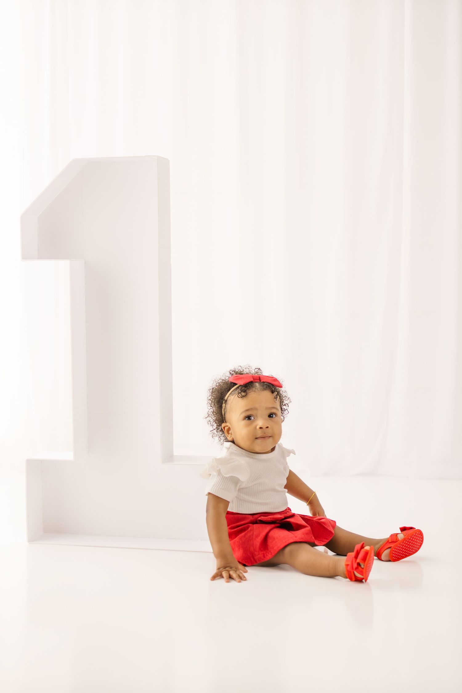 Baby girl in red outfit sits beside large number 1, celebrating her first birthday in a white studio.