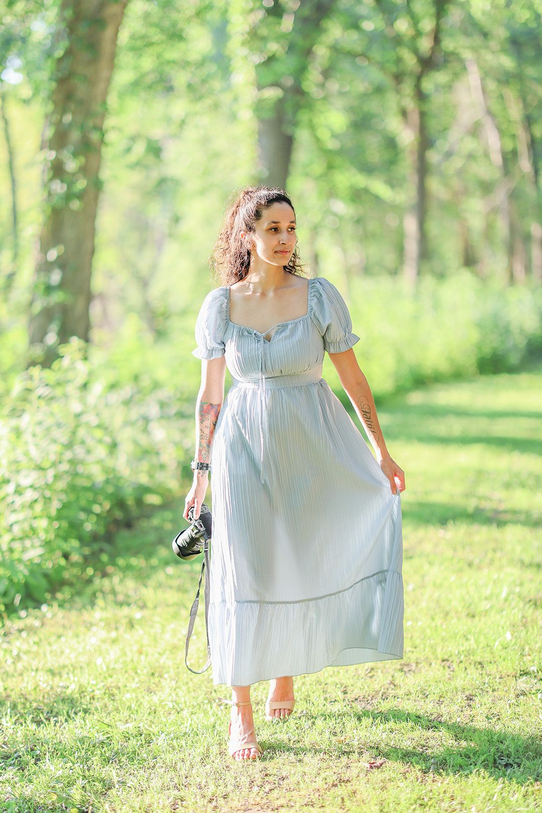 Woman in light blue midi dress holding camera walks through lush green park on sunny day.