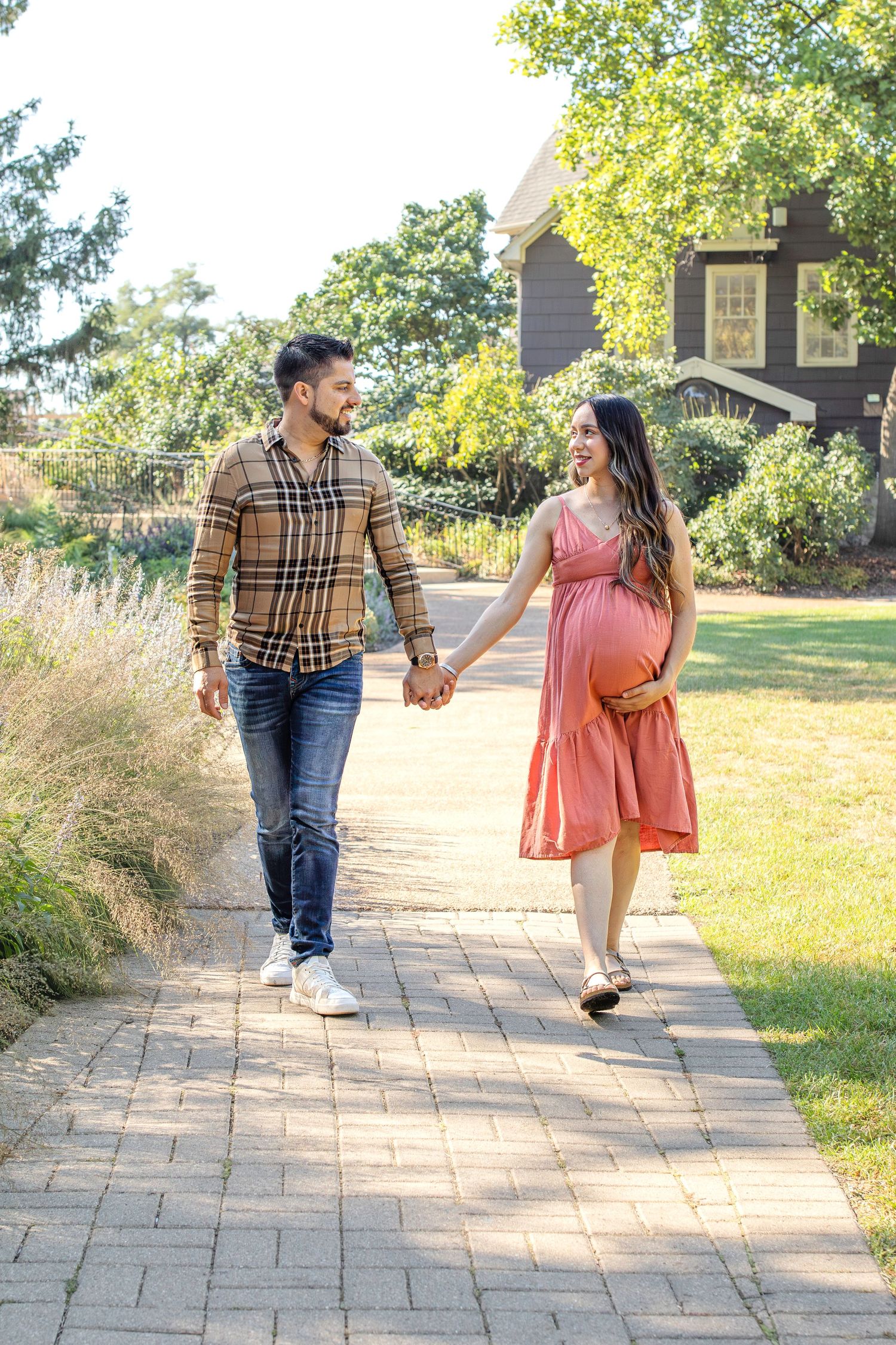 A couple walks hand in hand down a curved stone pathway surrounded by green grass and trees on a sunny day.