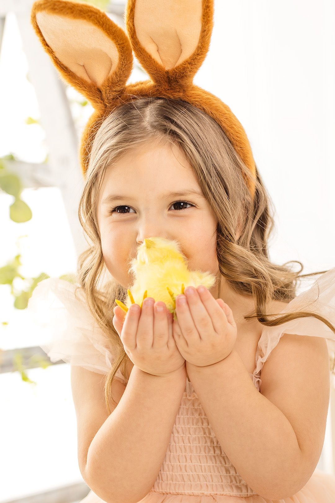 Child wearing bunny ears holds yellow chick while playing in bright sunlit room.