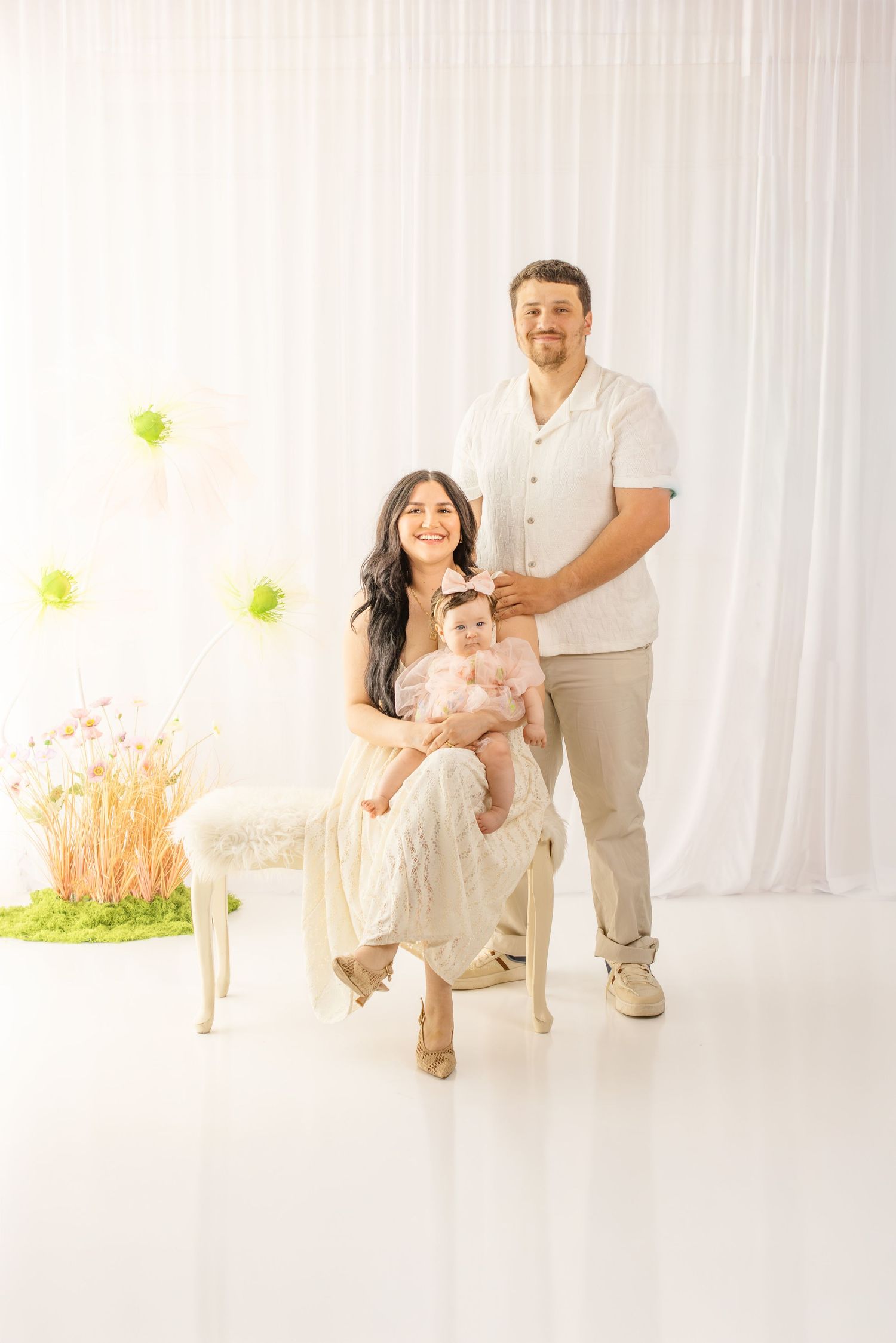 Family portrait of smiling parents with baby in white studio with floral decor and balloon backdrop.