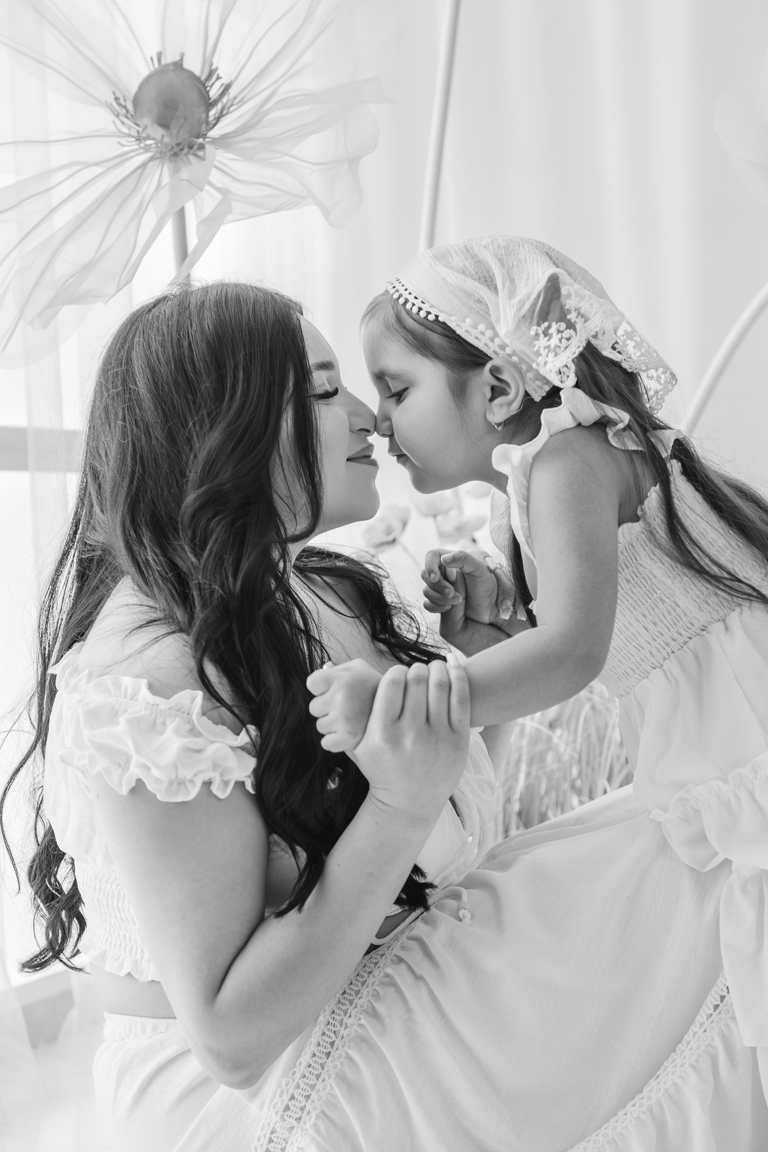 Black and white photo of a mother in white dress tenderly holding her young daughter close, touching noses.