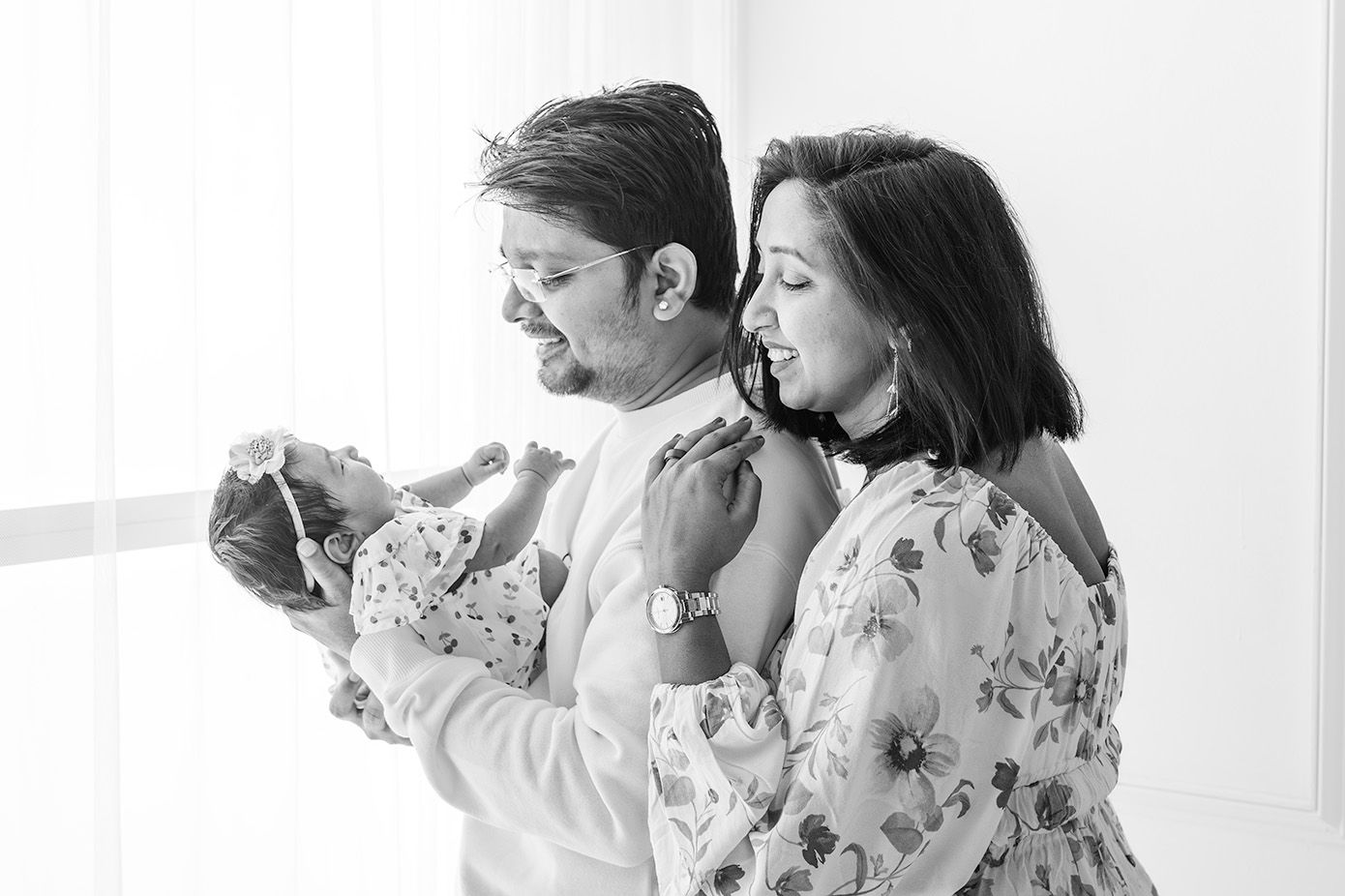 Black and white photo of a couple admiring their newborn baby near a bright window.
