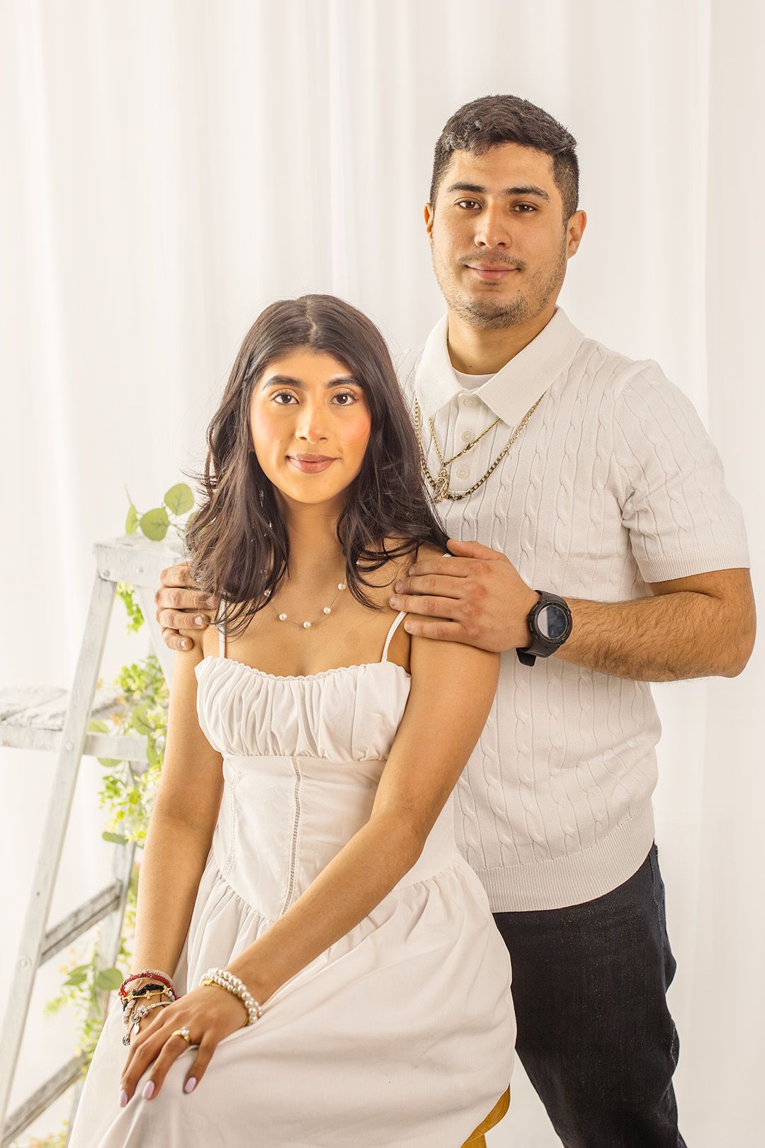 A young couple poses together in white outfits against a white curtain backdrop in a professional portrait photo.