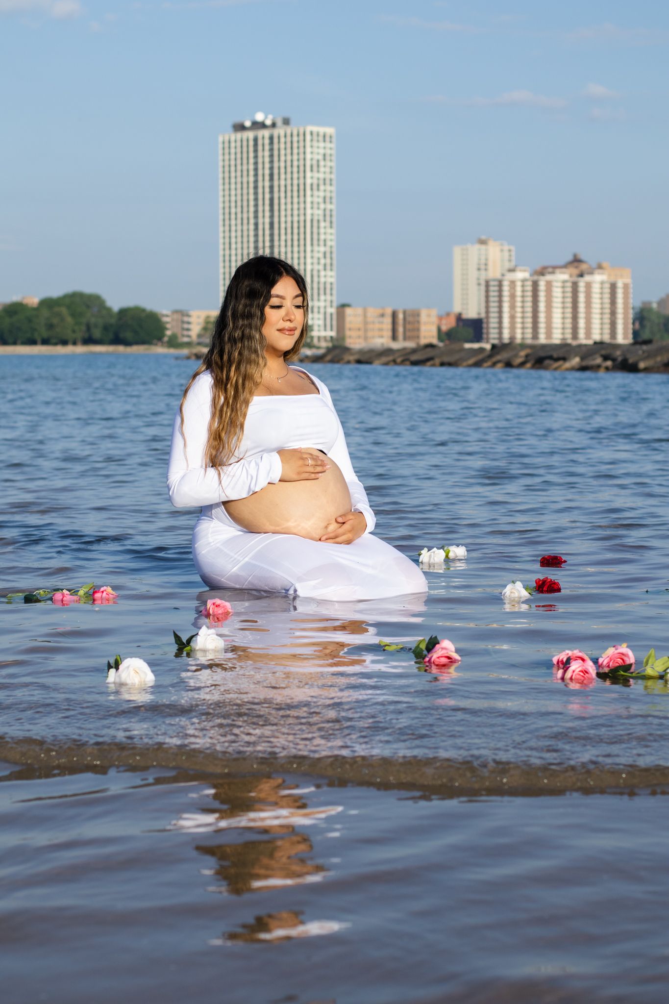 Pregnant woman in white sits in shallow water with floating flowers and cityscape behind.