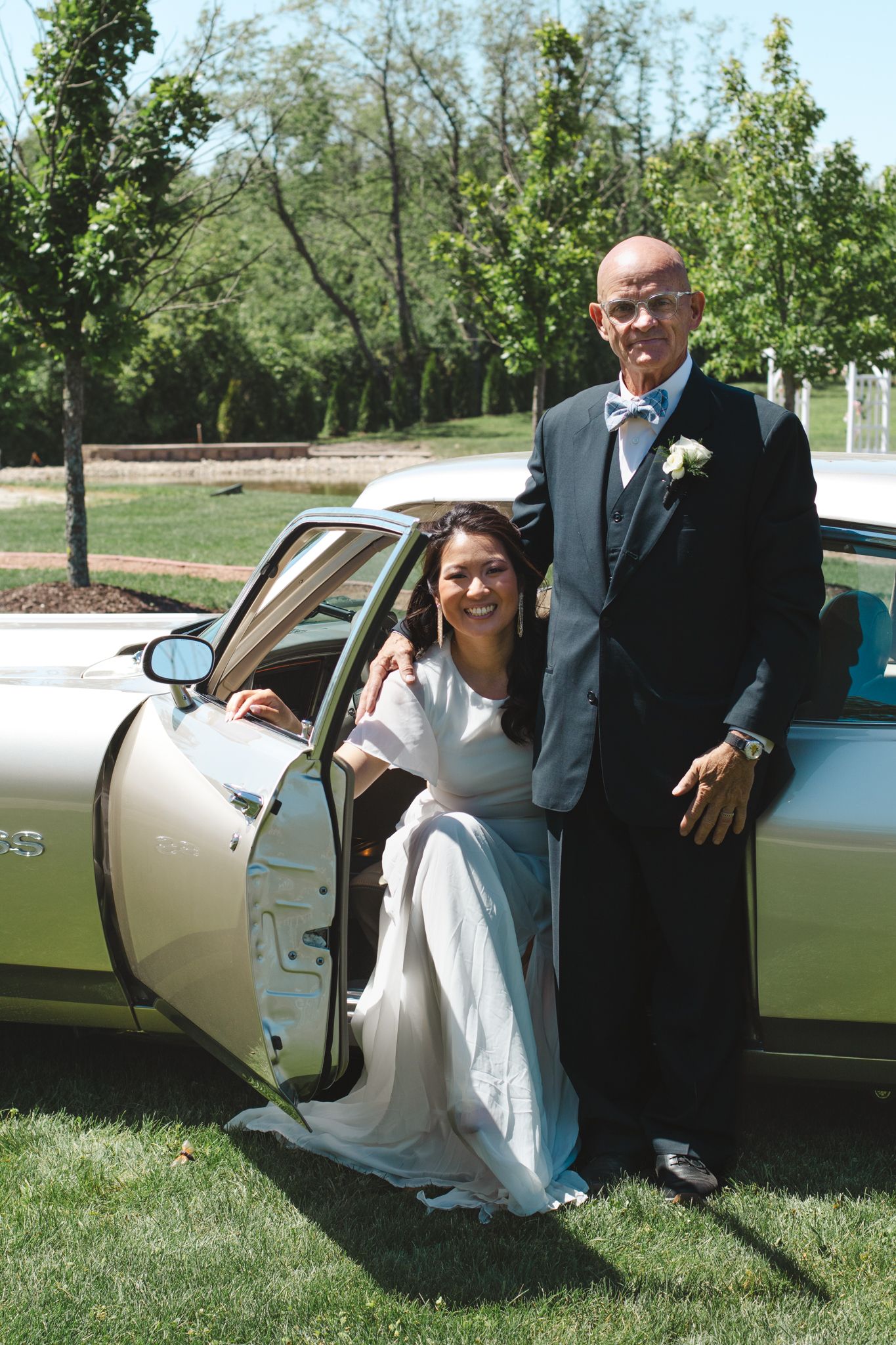 Dressed-up wedding attendees posing beside vintage champagne-colored car outdoors.