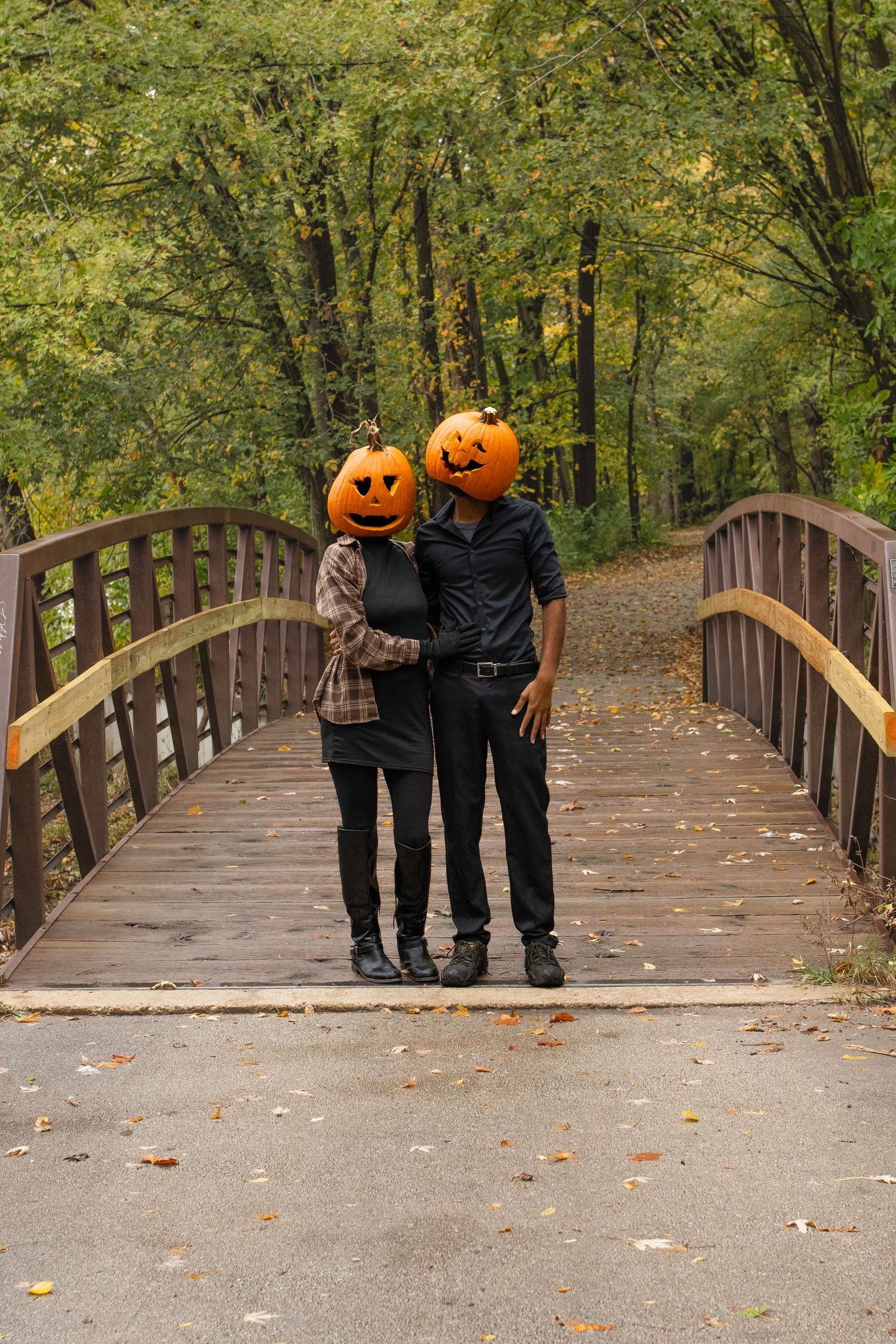 Two people holding pumpkins walk across a wooden bridge surrounded by autumn trees in a park setting.