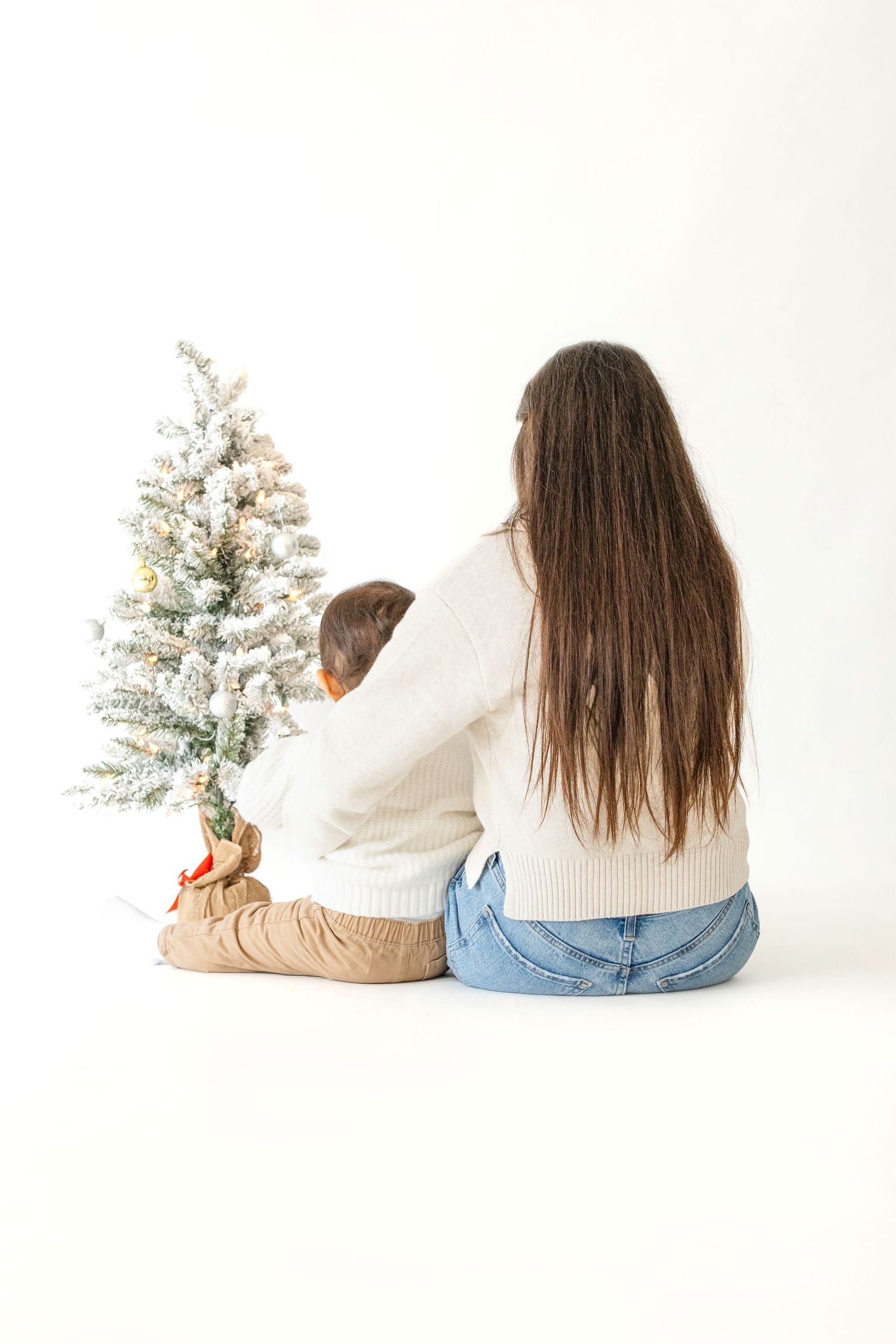 Two people in cozy sweaters sit beside a small flocked Christmas tree on white background.
