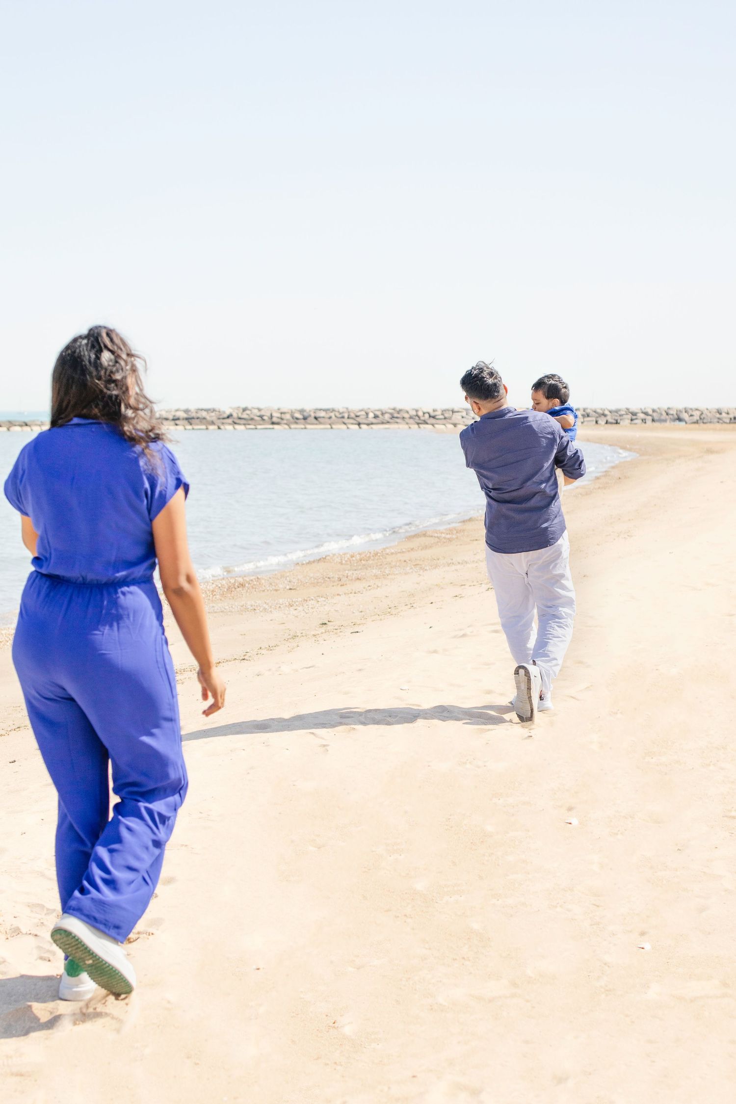 People walking and jogging on a sunny beach with a rocky breakwater visible in the background.